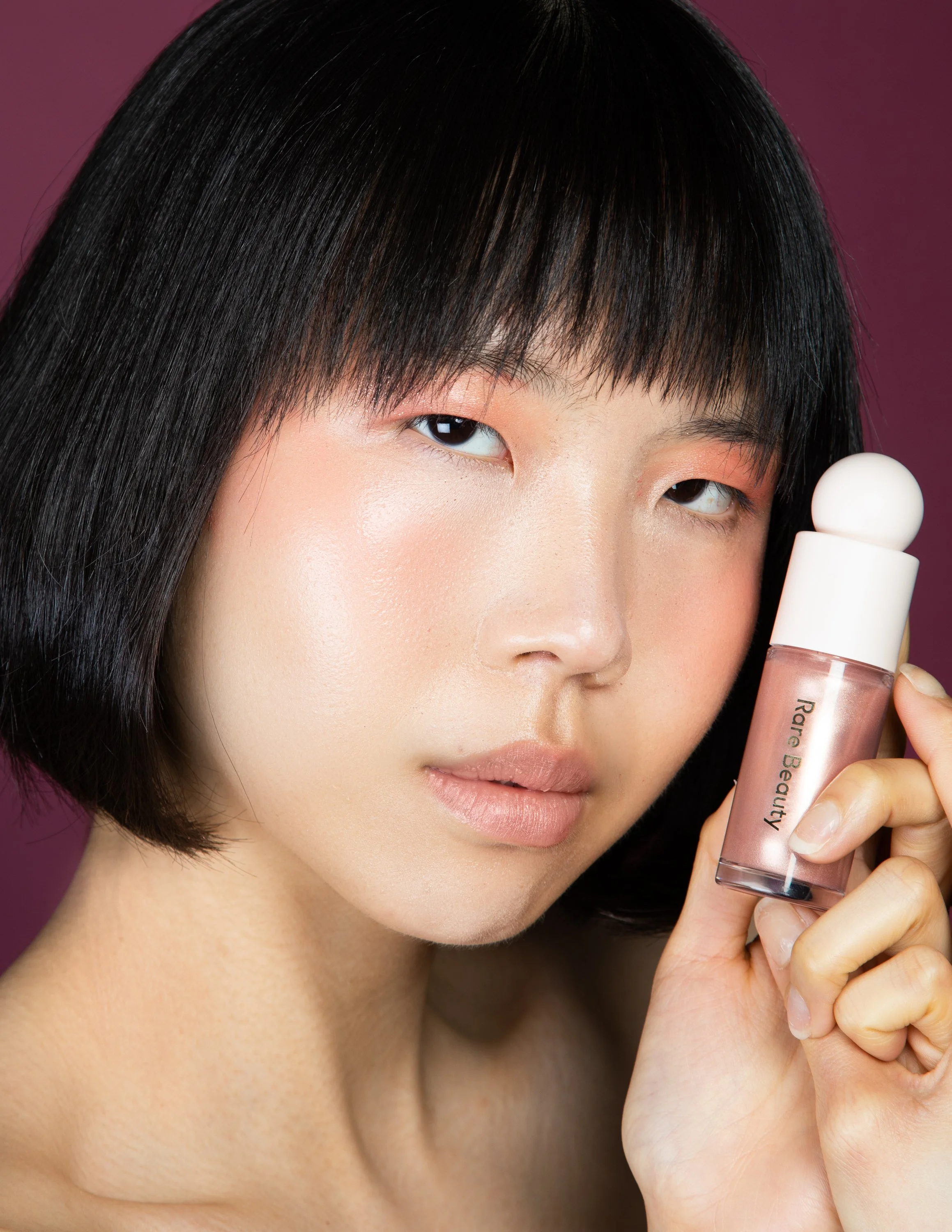 A woman with short black hair and natural makeup holding a bottle of Bare Beauty cosmetic product close to her face against a purple background.