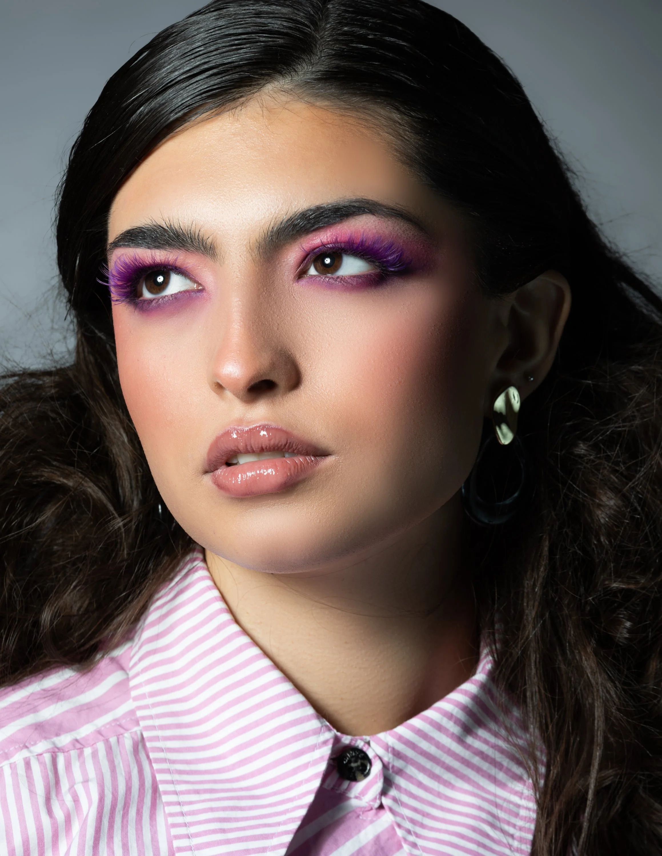 Close-up of a woman with dark hair, wearing purple eyeshadow, pink lipstick, and a pink and white striped collared shirt.