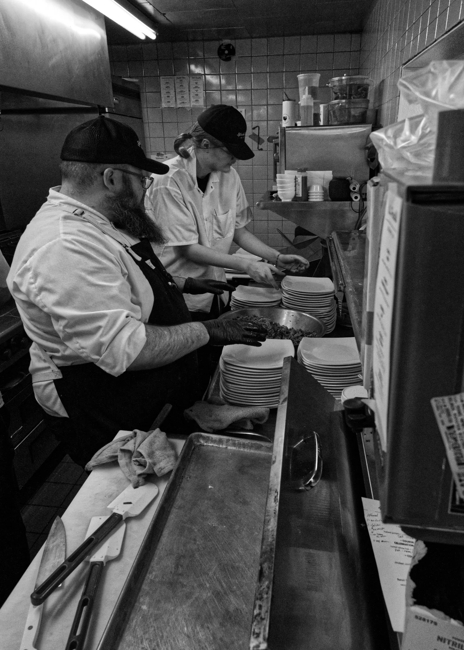 Two chefs in a commercial kitchen preparing food, one chopping or mixing ingredients while the other arranges plates.