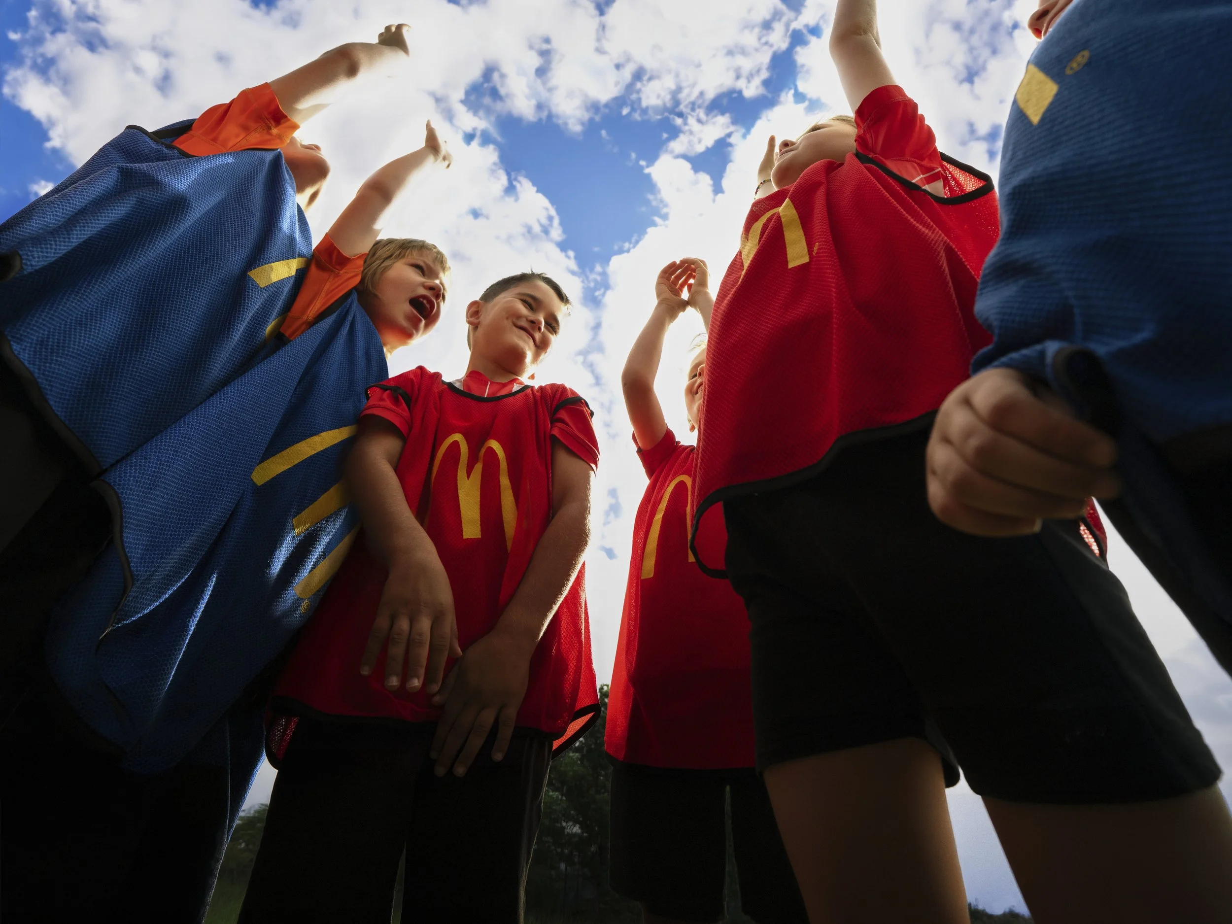 Group of children in colorful sports jerseys standing in a circle, with their arms raised towards the sky, looking at each other and smiling under a partly cloudy sky.