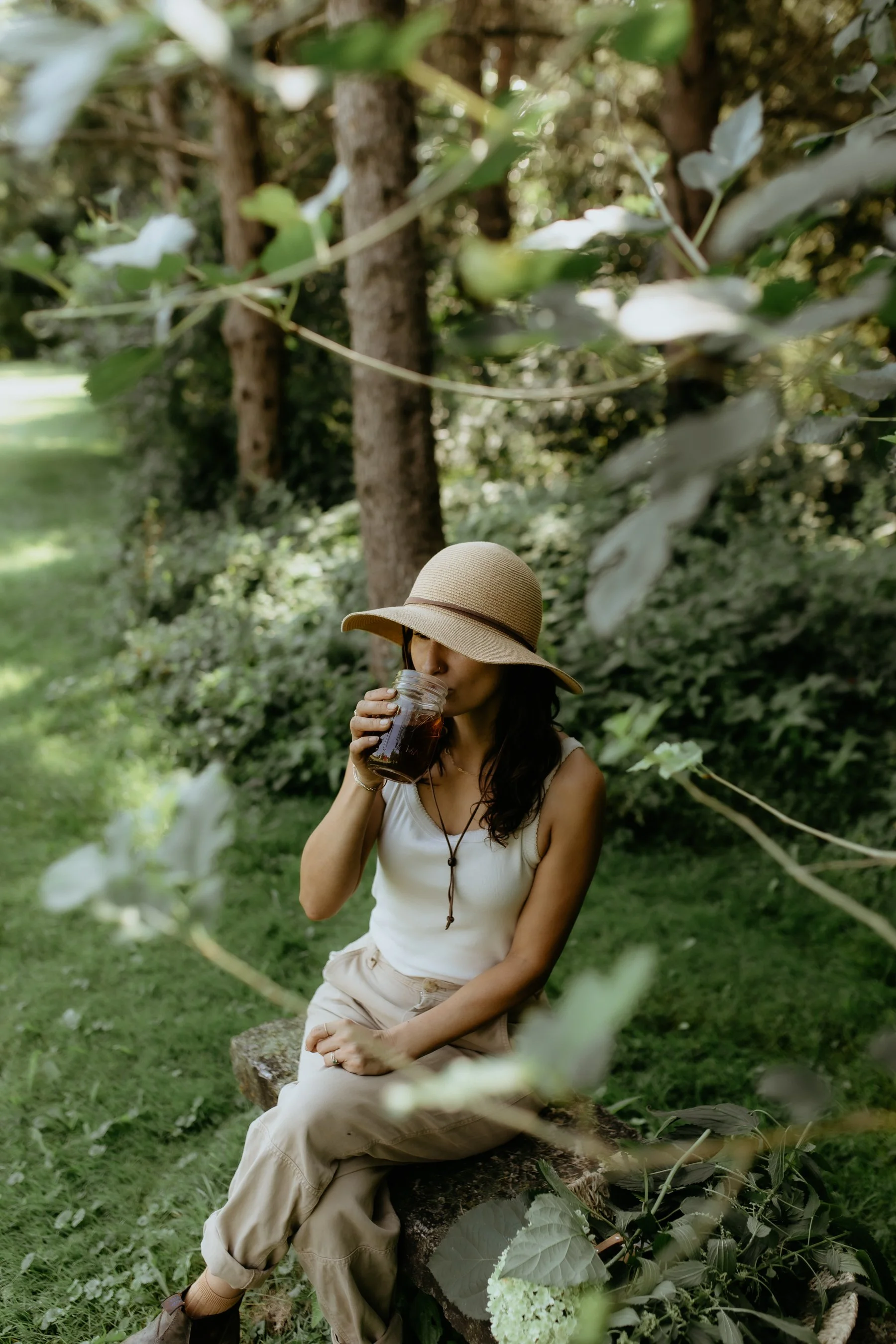 Herbalist and Founder of Growing Up Rooted, Daniela Rey, sitting on a log outdoors in a green, leafy area, wearing a wide-brimmed hat and drinking an herbal infusion from a jar. She is dressed in light-colored pants and a sleeveless top.
