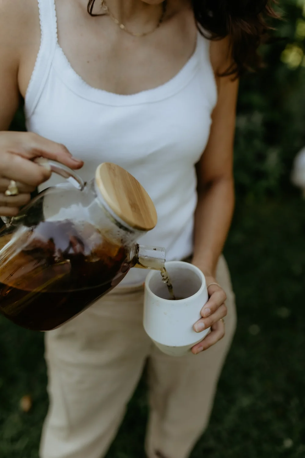 Daniela pouring an herbal infusion from a glass teapot into a mug in her garden