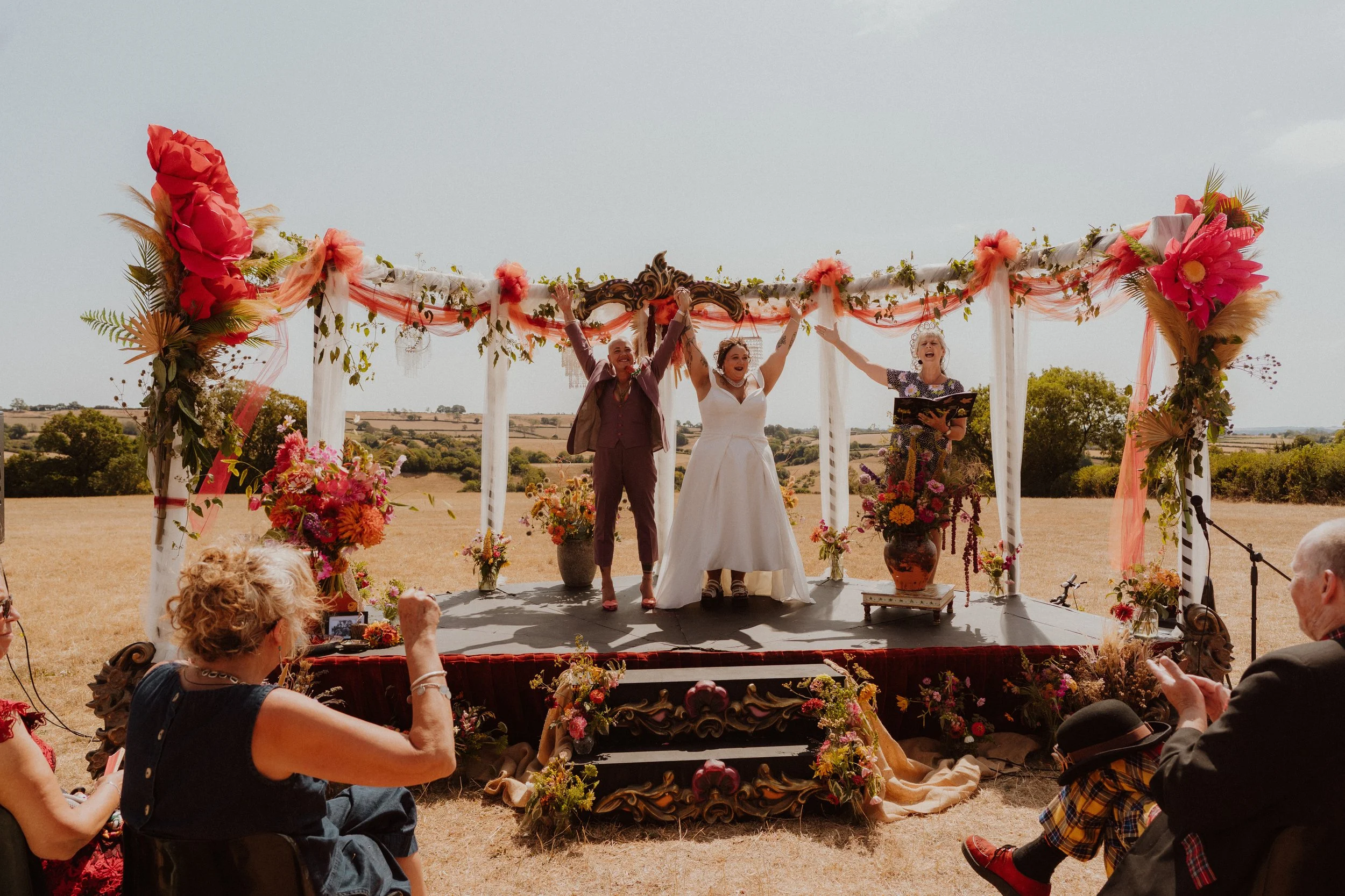 A wedding ceremony taking place outdoors under a decorated arch with three people celebrating, one holding a framed certificate, in a scenic rural landscape.