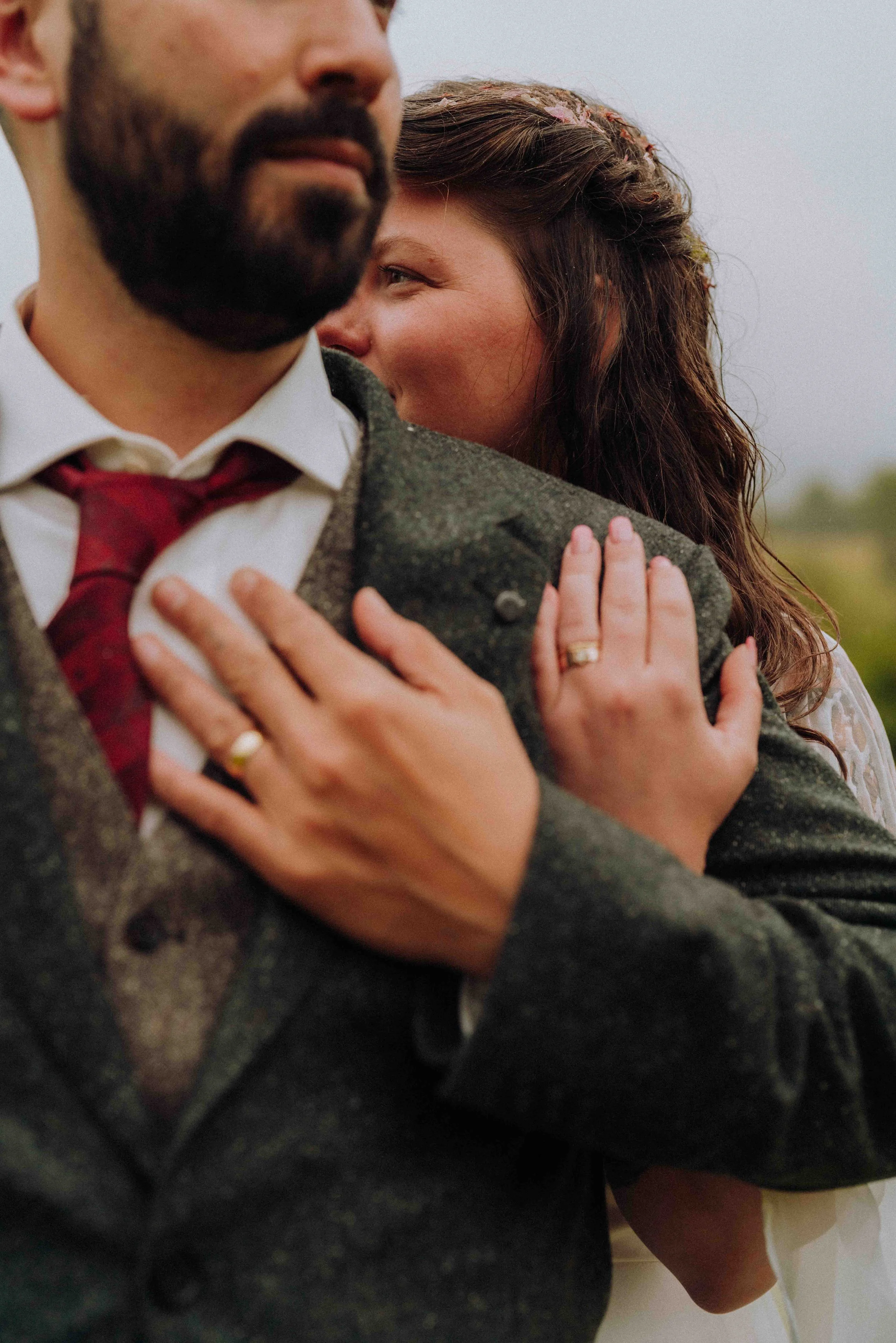 A close-up of a bride and groom hugging, with the man's face partially visible and the woman's face visible, smiling and showing a wedding ring on her finger.