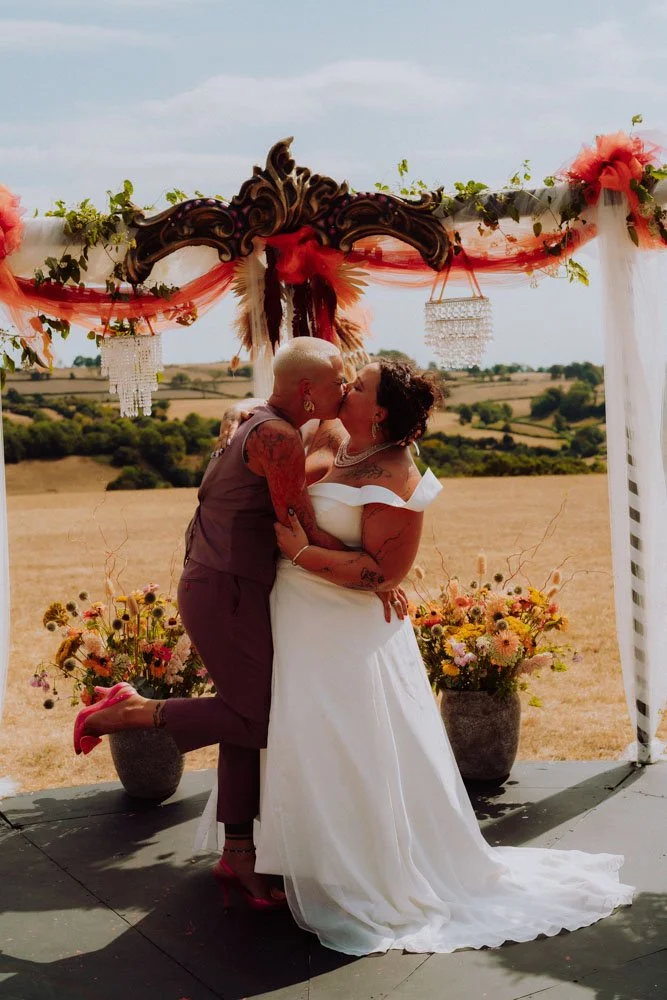 Merriers kissing at an outdoor wedding ceremony. One woman is in a white wedding dress, and the other partner is in a purple jumpsuit. Behind them is a decorated wooden arch with flowers and greenery, with an open field and hills in the background.