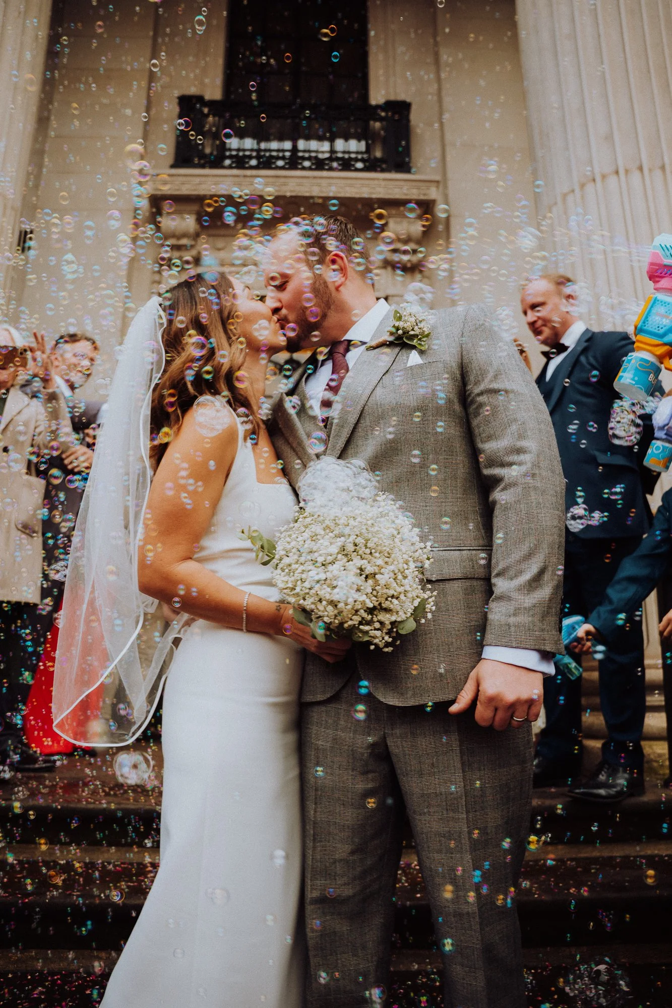 newly weds having a kiss on the stairs of the Old Marlybone Town Hall in London whilst their guests are blowing bubbles on them