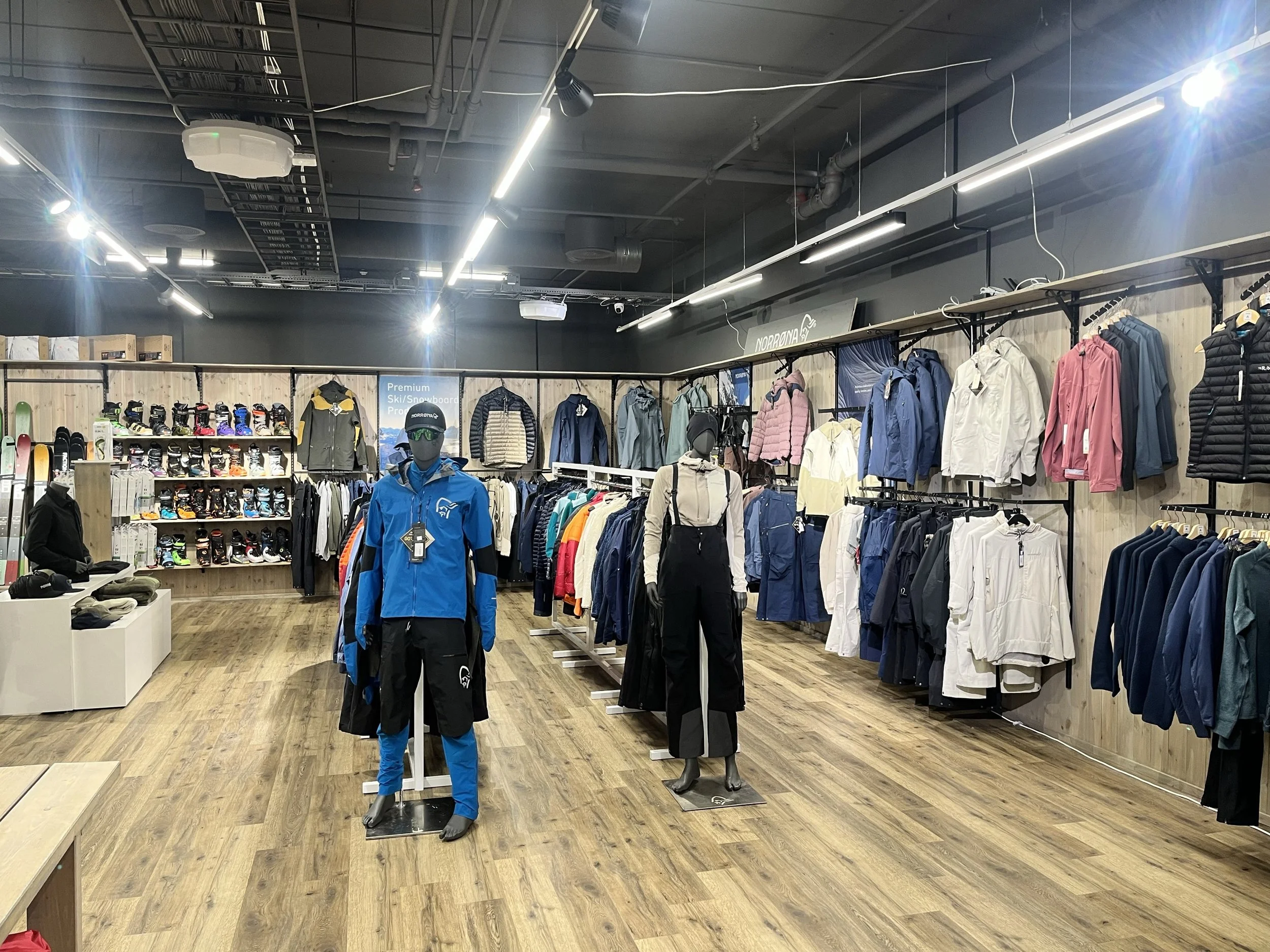 Herre- og dametøy samt skiklær og sportsutstyr utstilt i en sportsbutikk med tre-parkett og moderne belysning.