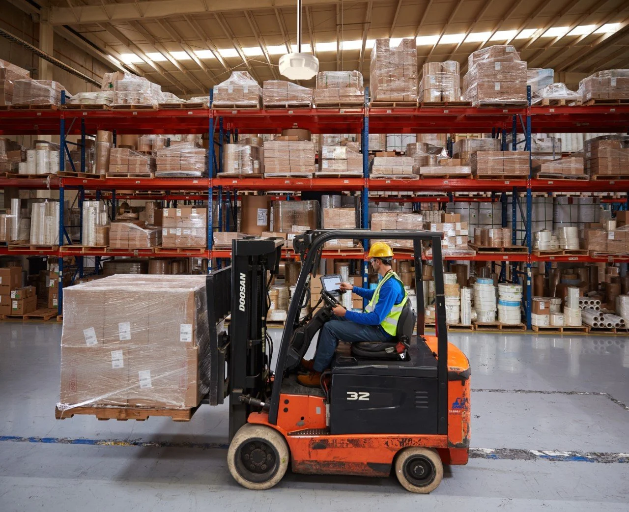 En person i en lagerhall kjører en liten gaffeltruck med en stor, pakket palle.