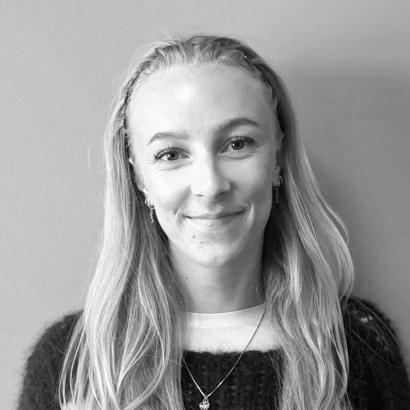 Sort-hvit foto av en smilende kvinne med langt, blond hår, iført mørk genser og smykker, mot en enkel vegg.