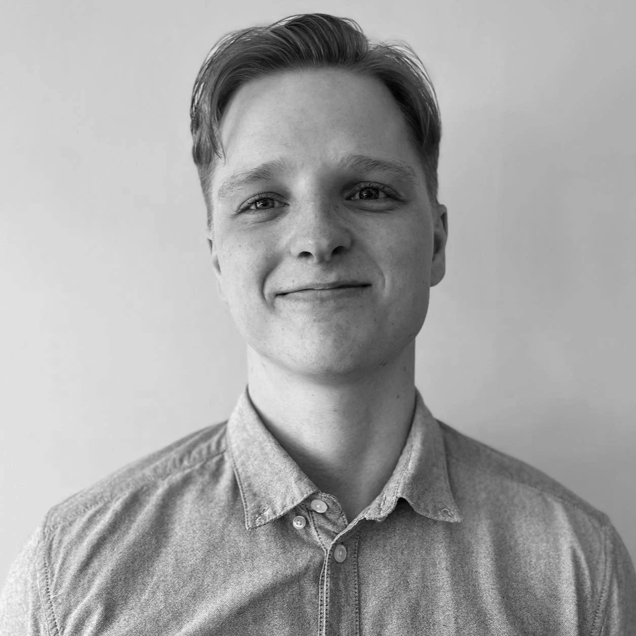 Svart-hvitt portrett av en smilende ung mann med kort, lyst hår, iført en knappet skjorte, mot en enkel lys bakgrunn.