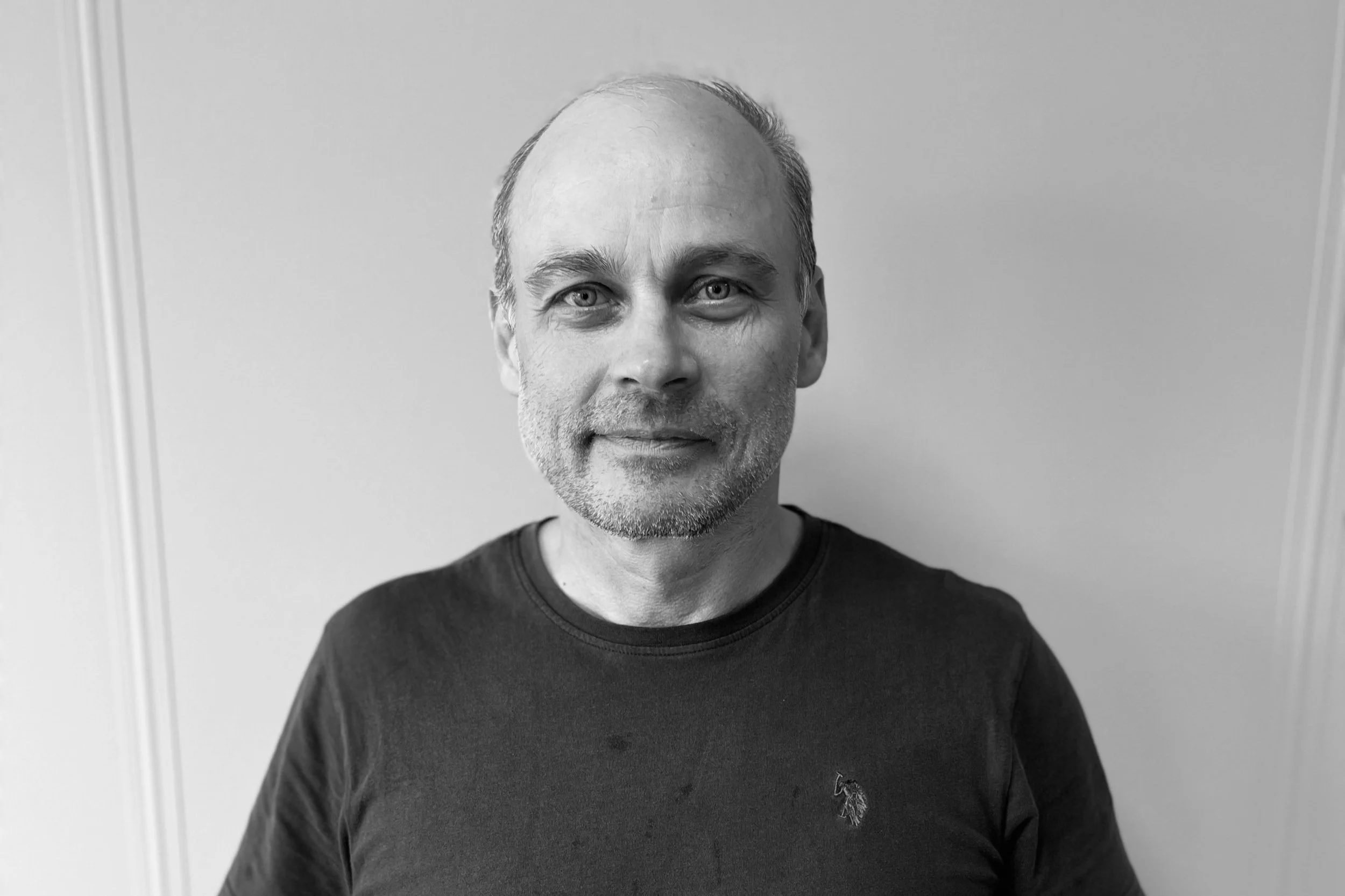 Portrett av en mann med lyst hår og skjegg, iført svart t-skjorte, stående foran en enkel vegg