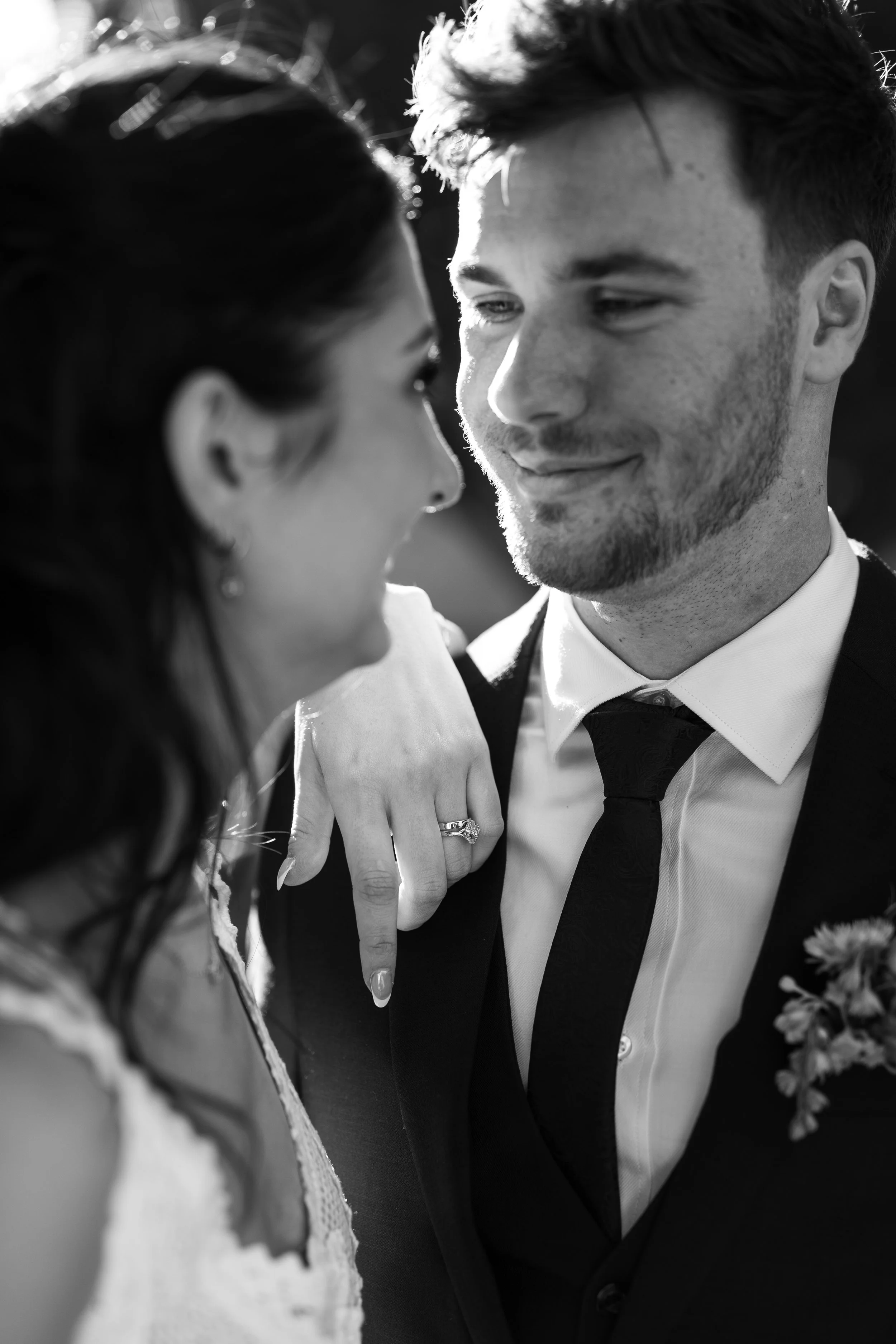 A black-and-white photo of a couple close together, smiling and looking at each other, with the woman's hand resting on the man's shoulder showing her engagement ring.