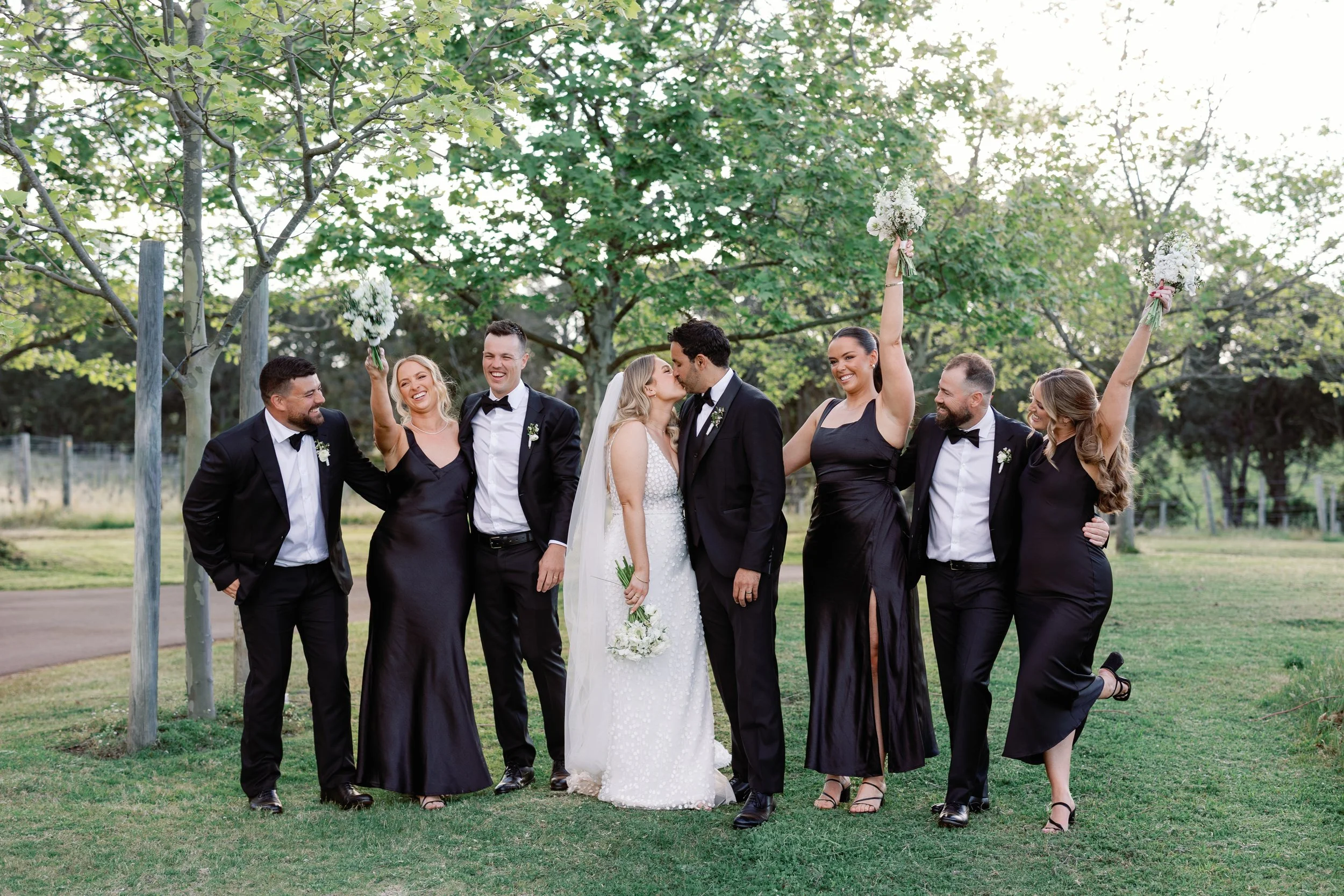 A wedding party of seven people standing outdoors on grass under trees, celebrating, with two bridesmaids holding bouquets with pink ribbons raised in the air, and the bride and groom kissing in the center.