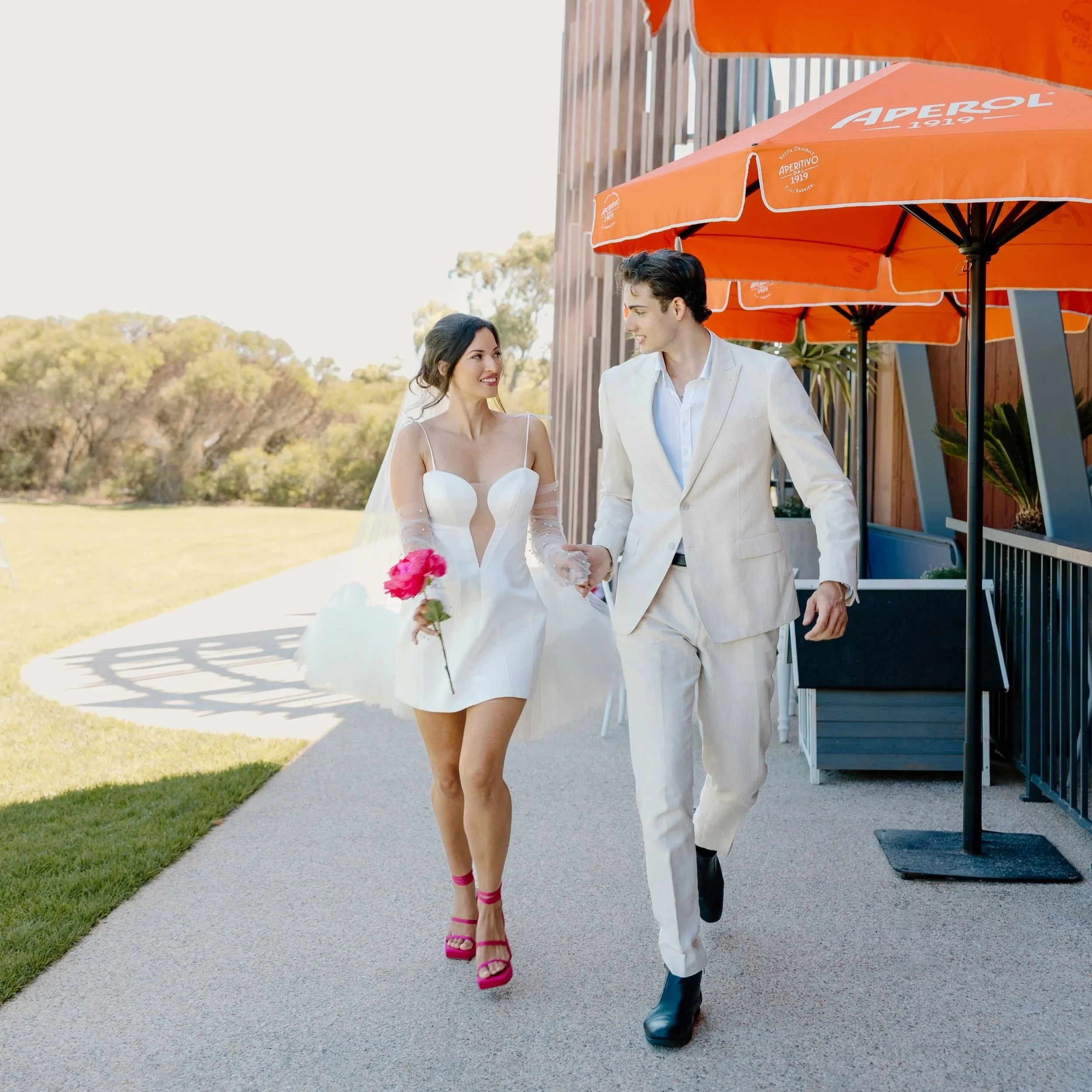 A bride and groom walking outdoors, holding hands and looking at each other. The bride wears a white dress with a keyhole neckline and pink heels, holding a pink flower. The groom wears a white suit with black boots. They walk on a paved path near or