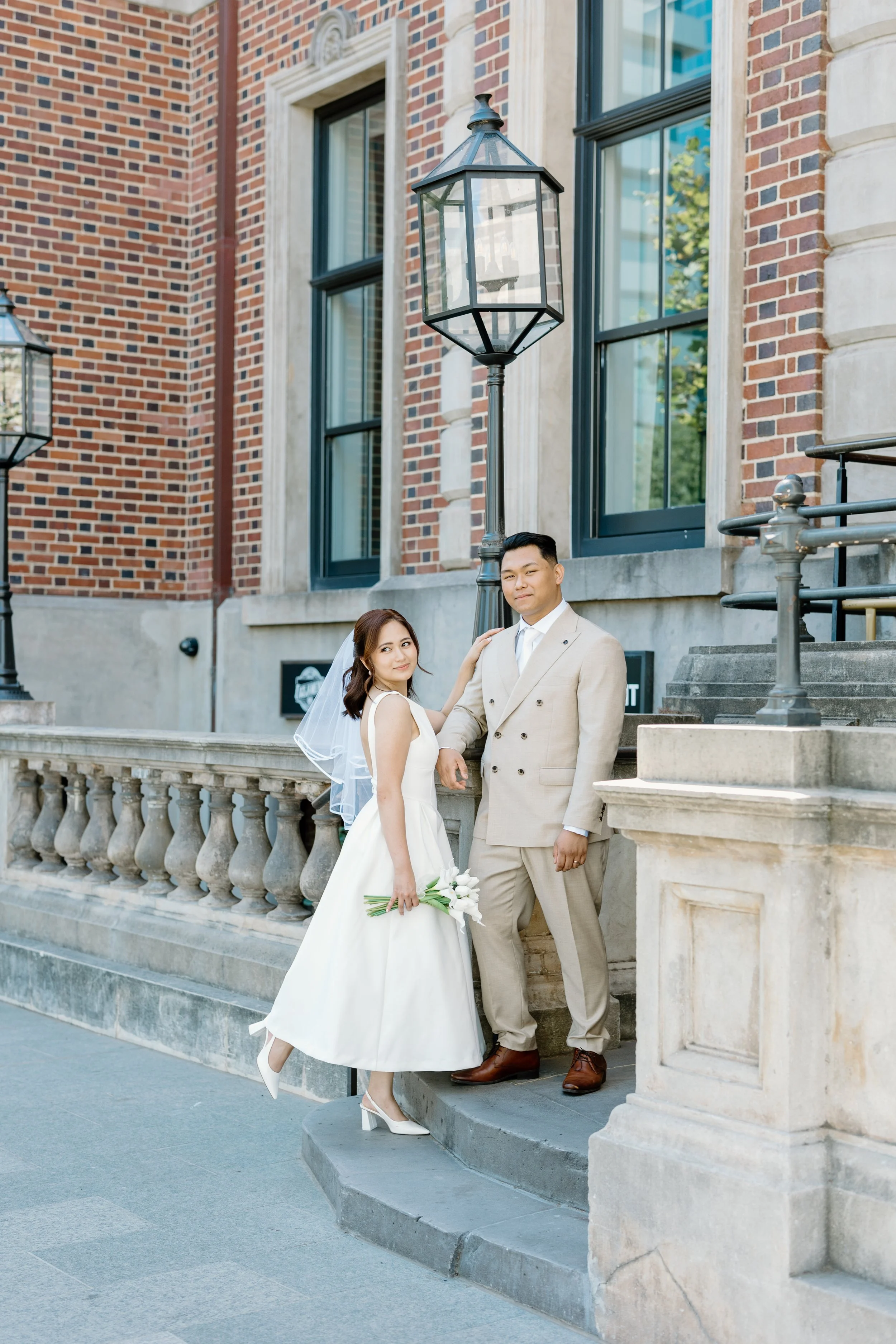 A bride and groom posing outside a brick building with large windows and vintage street lamps, the bride holding a small bouquet of white flowers at the State Buildings Perth