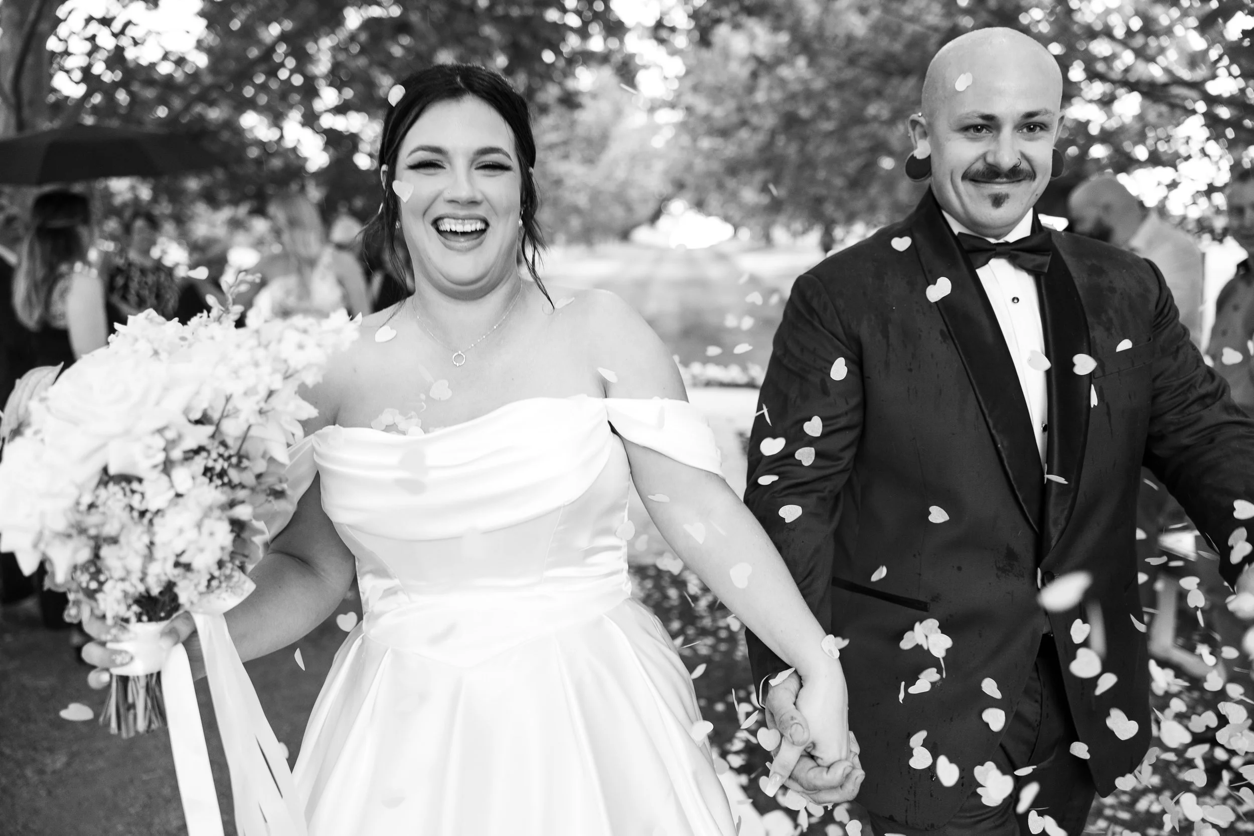 A happy bride and groom walking hand in hand during their wedding celebration, with confetti falling around them, outdoors with trees and guests in the background in South West WA
