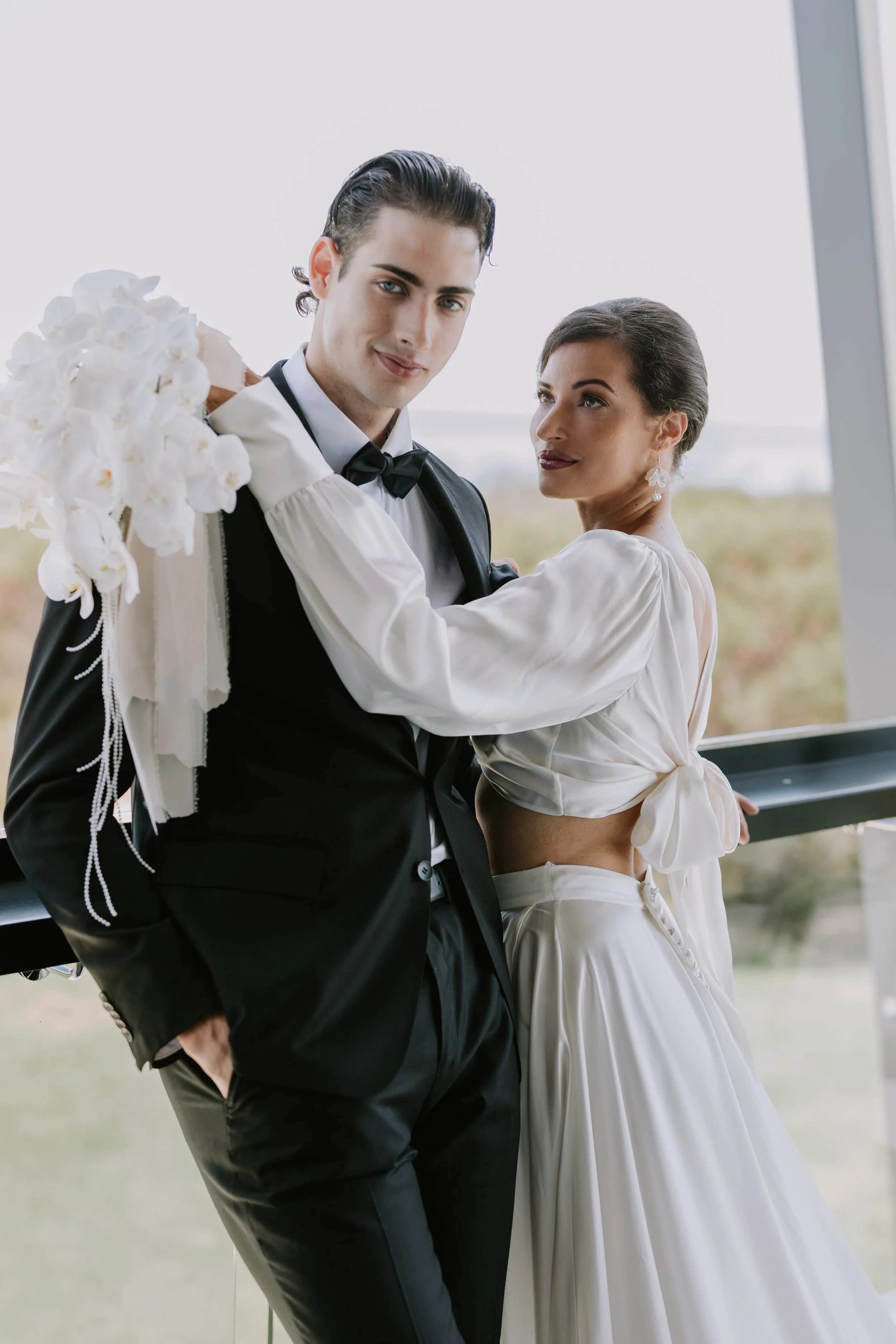 A couple, dressed in wedding attire, stands close to each other by a large window. The man wears a black tuxedo and bow tie, holding a bouquet of white flowers over his shoulder. The woman, in a white dress with a side bow, looks at the camera.