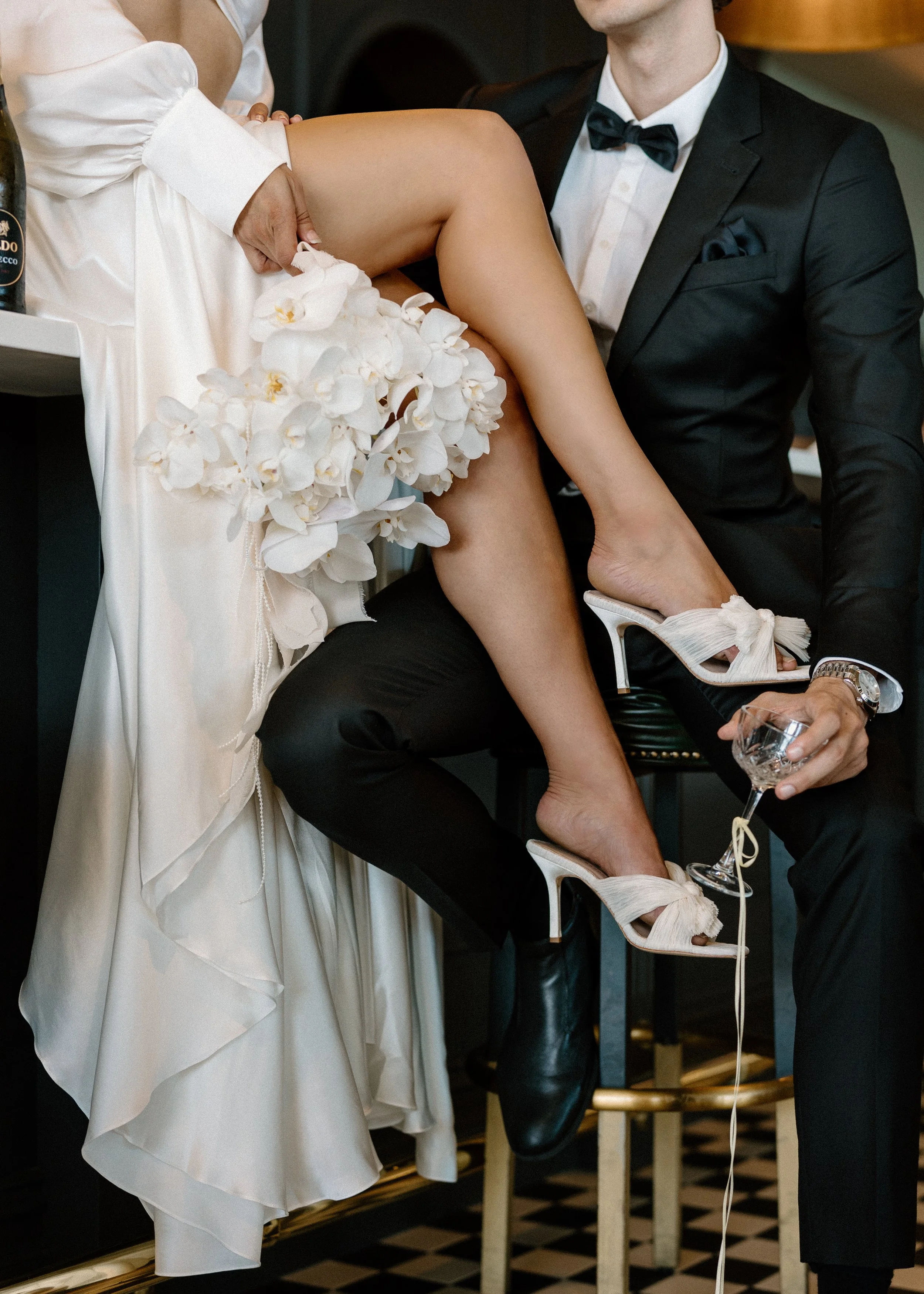 A woman in a white dress sitting on a man's lap. The woman is lifting her dress, revealing her leg and shoes, which are high heels with bows. The man is dressed in a black tuxedo with a bow tie, holding a glass and sitting on a chair.