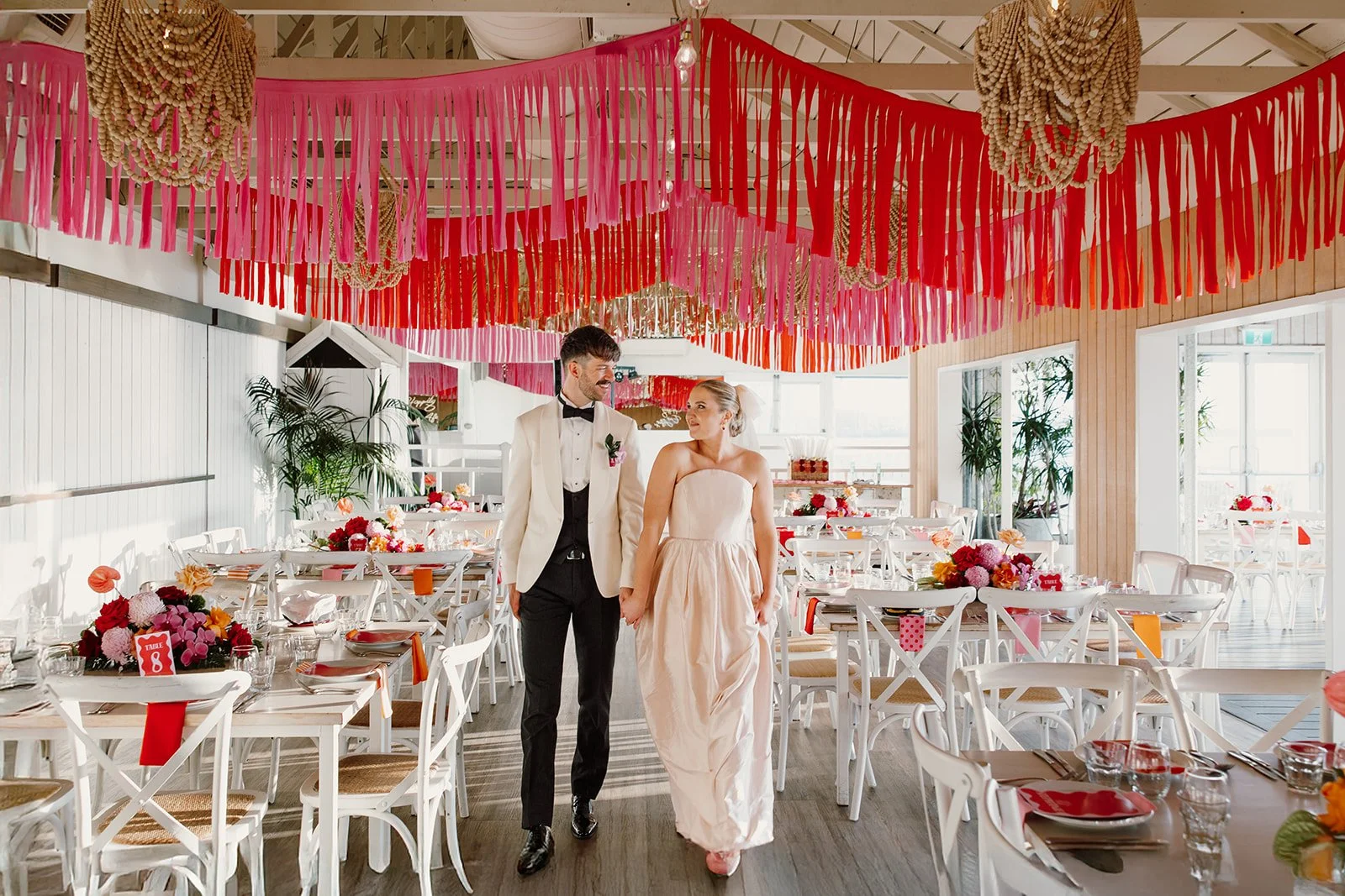 A bride and groom walking hand in hand through a decorated wedding reception hall with pink and red streamers and floral centerpieces.