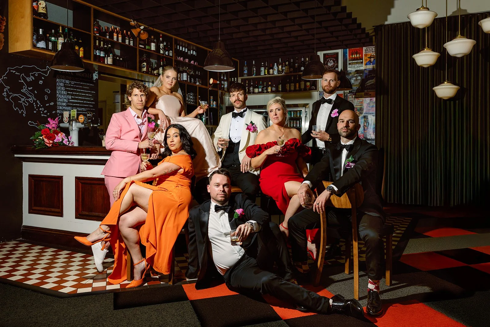 Group of eight people dressed in formal and semi-formal attire posing for a photo at a bar or restaurant, holding drinks, with shelves of bottles and posters in the background.