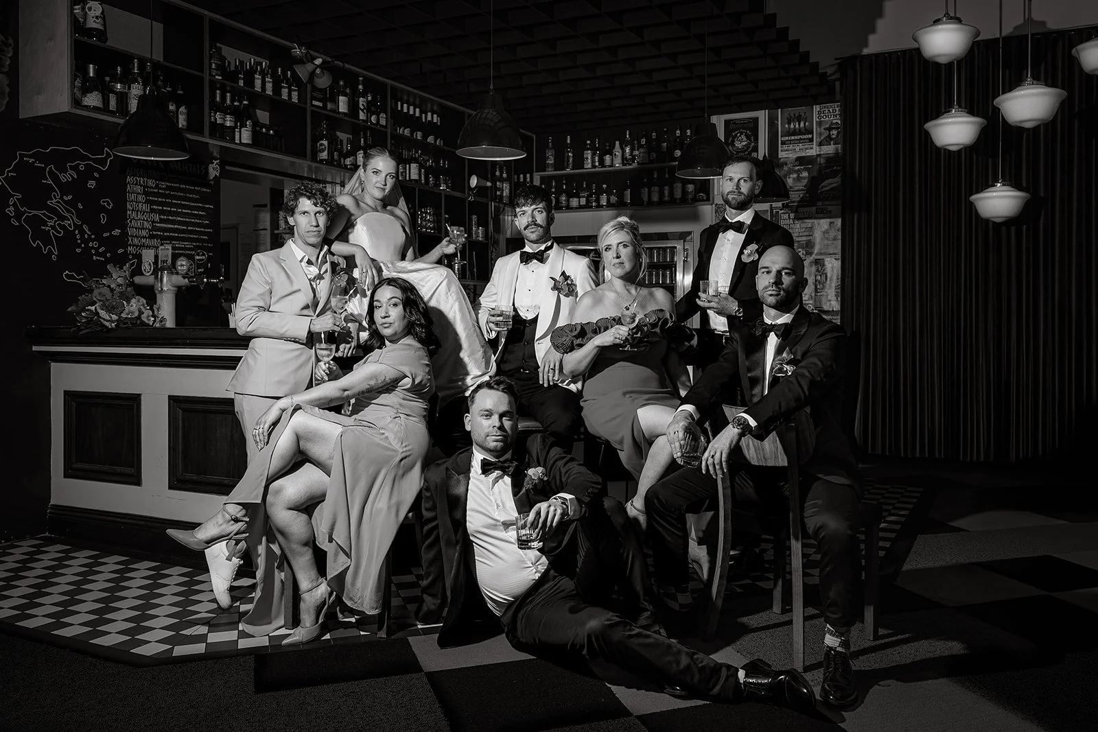 Group of people dressed in formal attire posing in a bar or lounge setting, some holding glasses of wine or cocktails, with shelves of bottles and bar decor in the background.