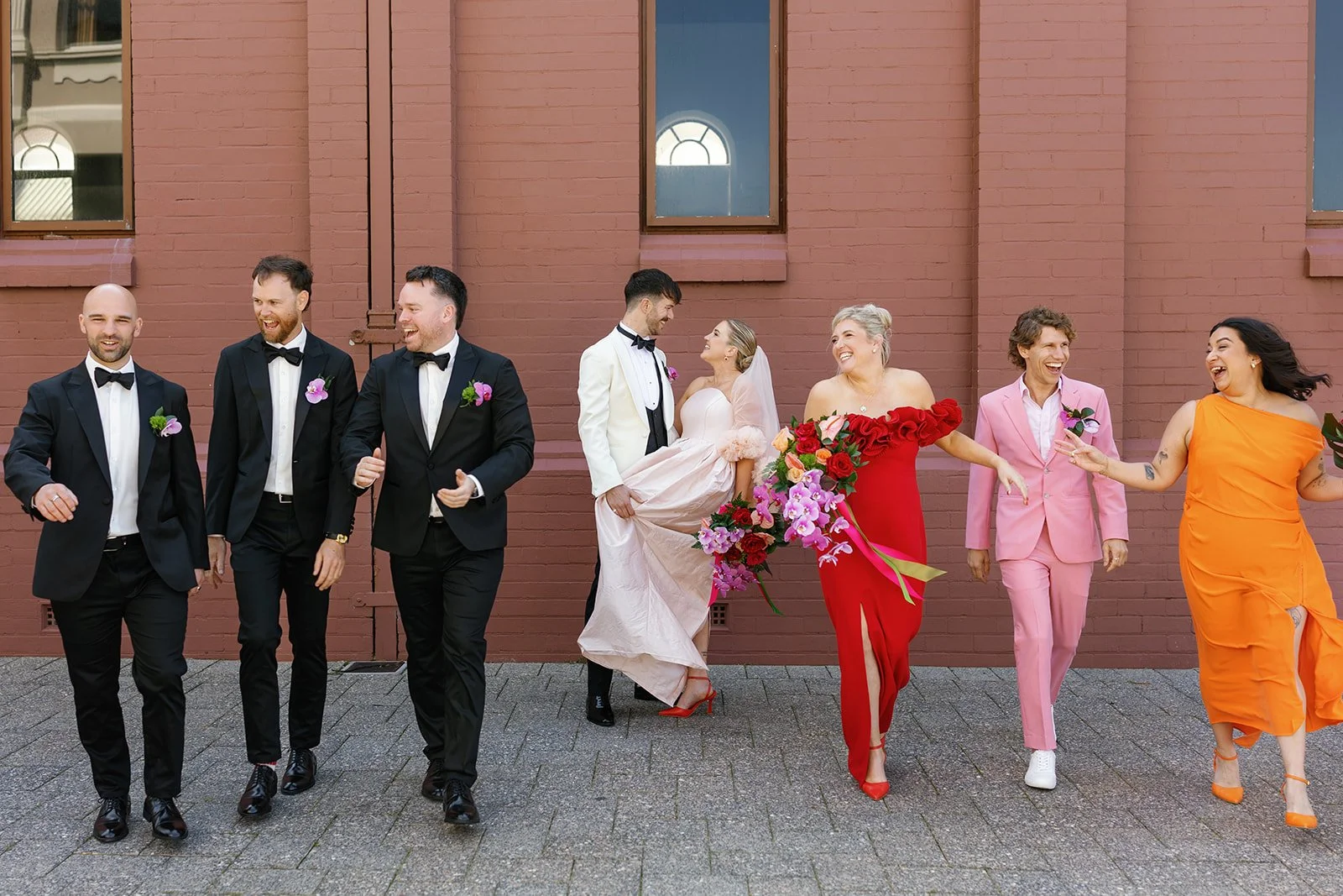 Group of seven people, including a bride and groom, walking and smiling in front of a brick wall. The bride is wearing a light pink wedding dress, and the groom is in a white tuxedo with black bow tie. Two women are dressed in bright red and orange d