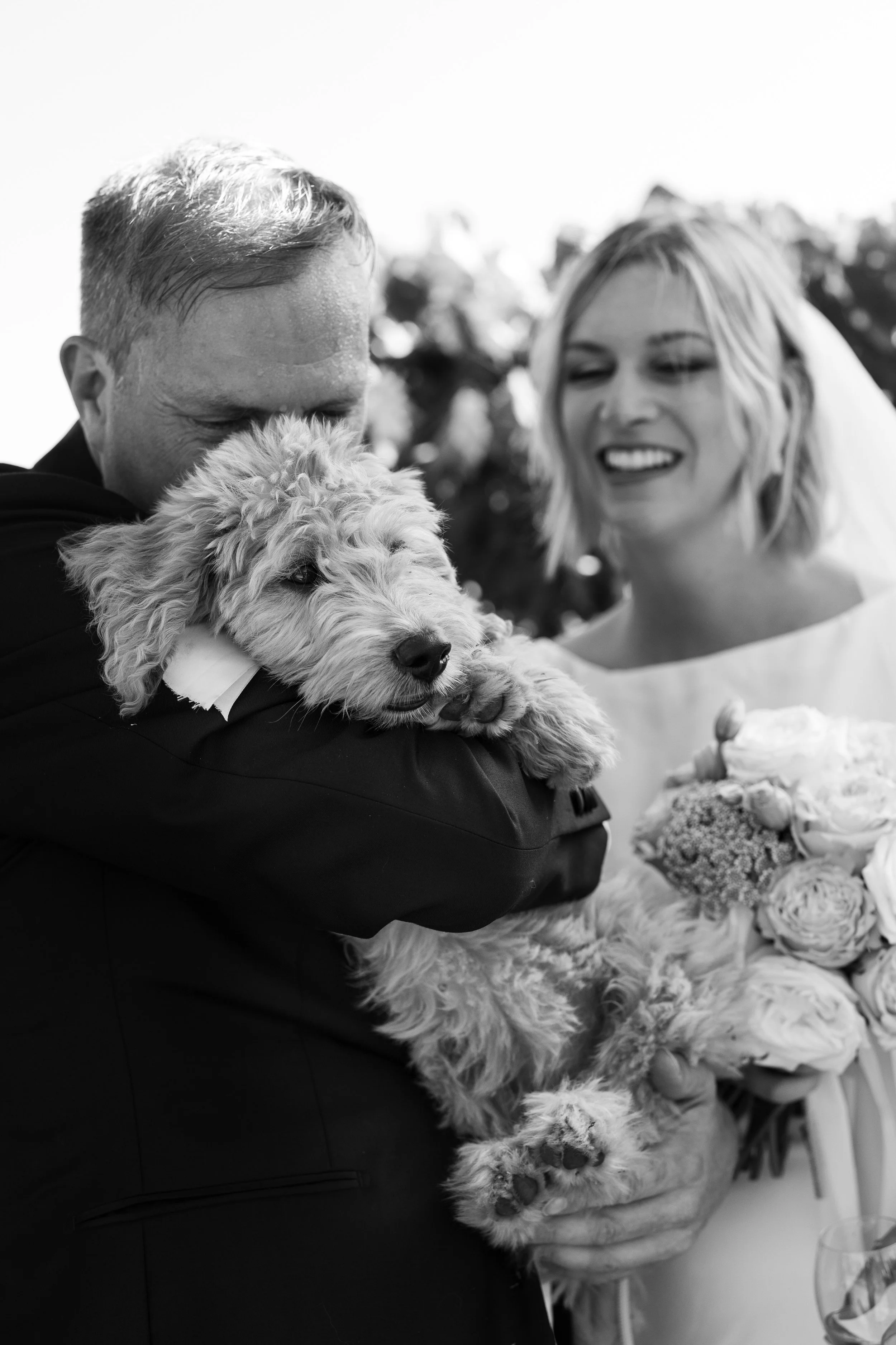 Black and white photo of a man and woman at a wedding, holding a small dog, with the woman holding a bouquet of flowers.