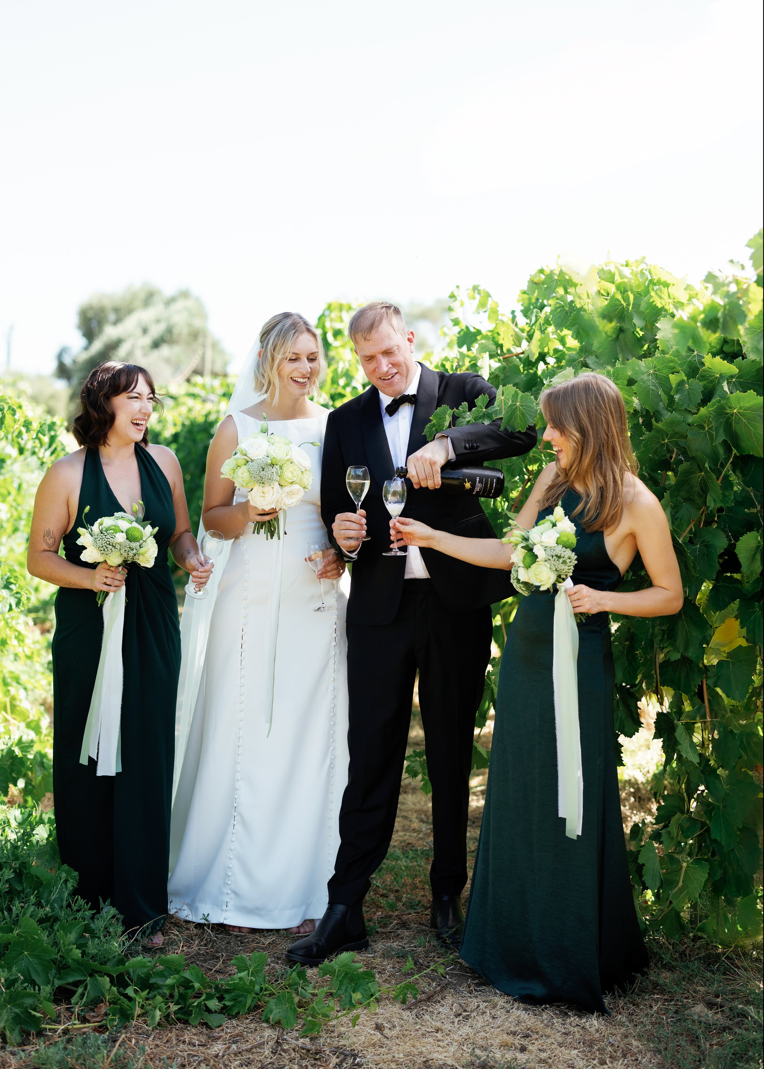 A wedding celebration outdoors with four women and one man, the man is pouring champagne into glasses held by the women, all are smiling and dressed in formal attire, surrounded by green foliage.