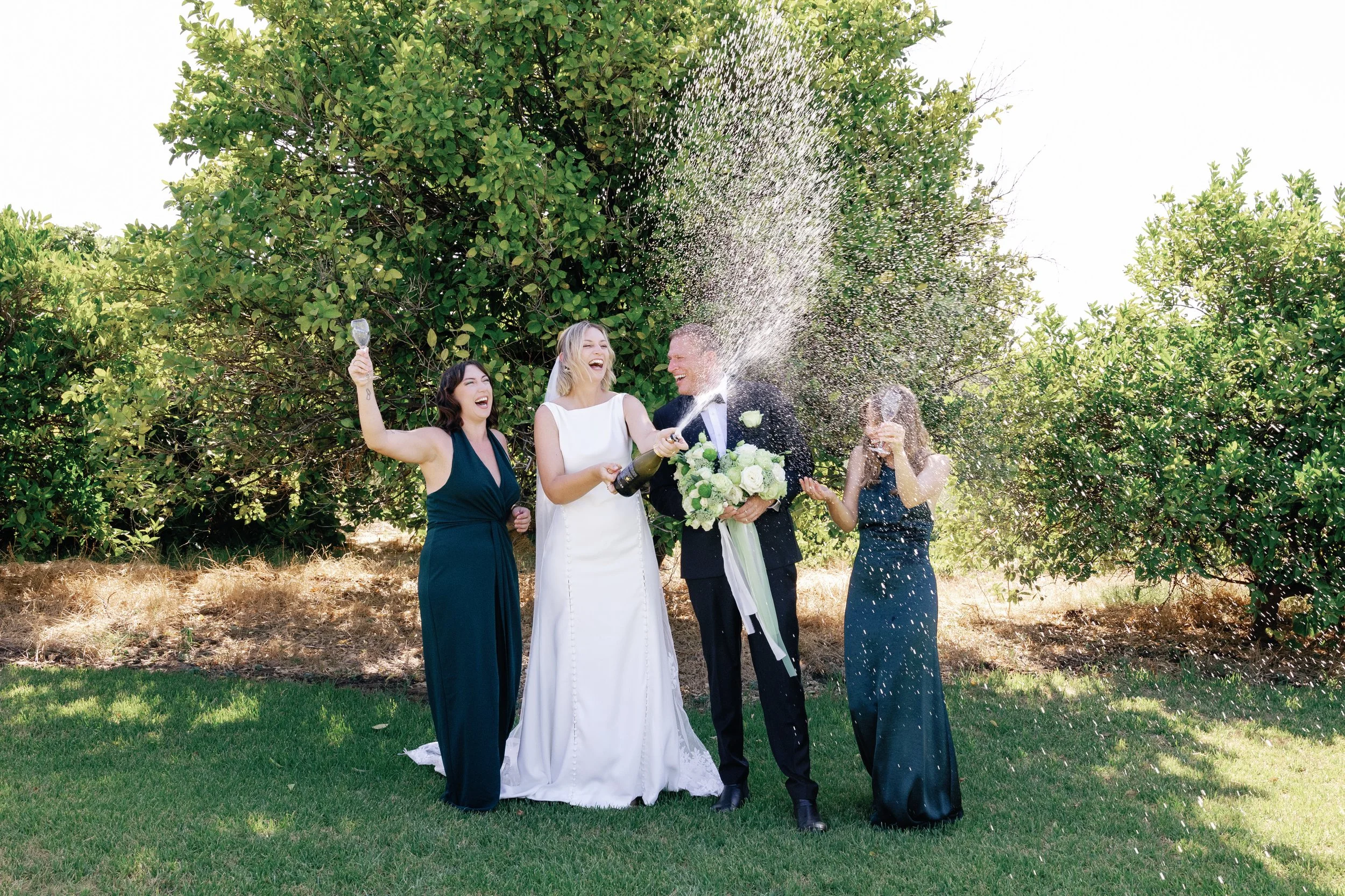 A wedding celebration outdoors with four women, one in a white wedding gown and holding a bouquet, and one man in a tuxedo. They are happily spraying champagne and celebrating under green trees on a sunny day.