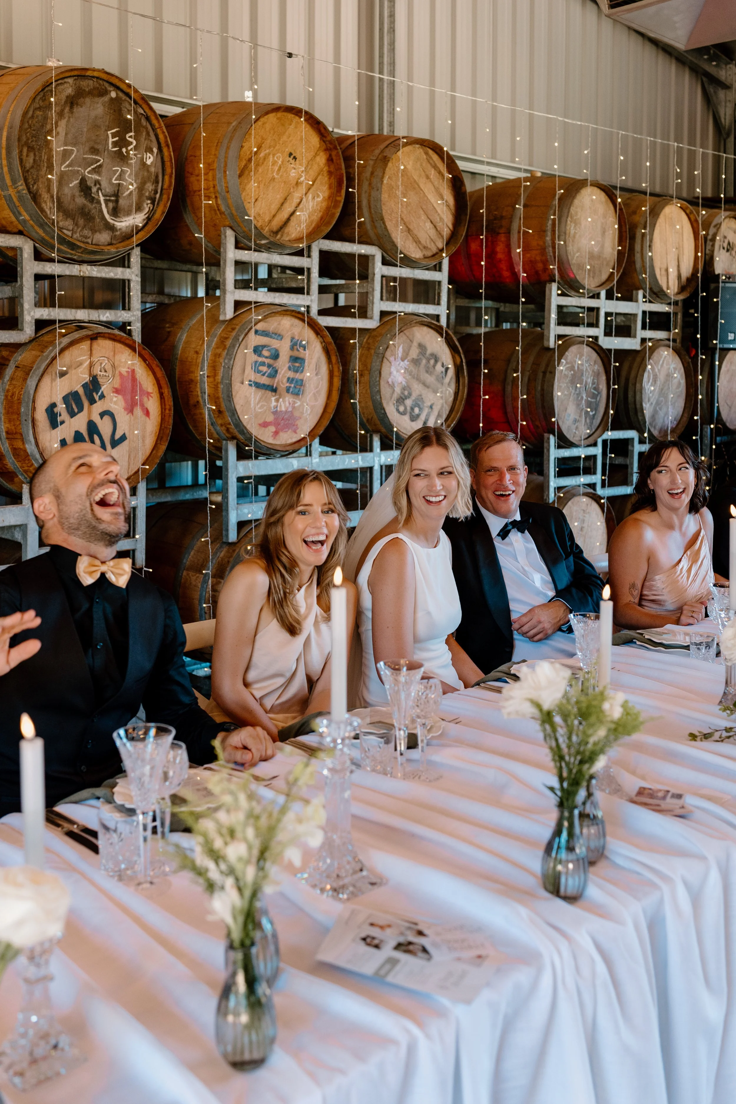 Group of people at a wedding reception, sitting at a long white table with floral centerpieces and candles, laughing and smiling, with wine barrels decorated with Canadian flags in the background.