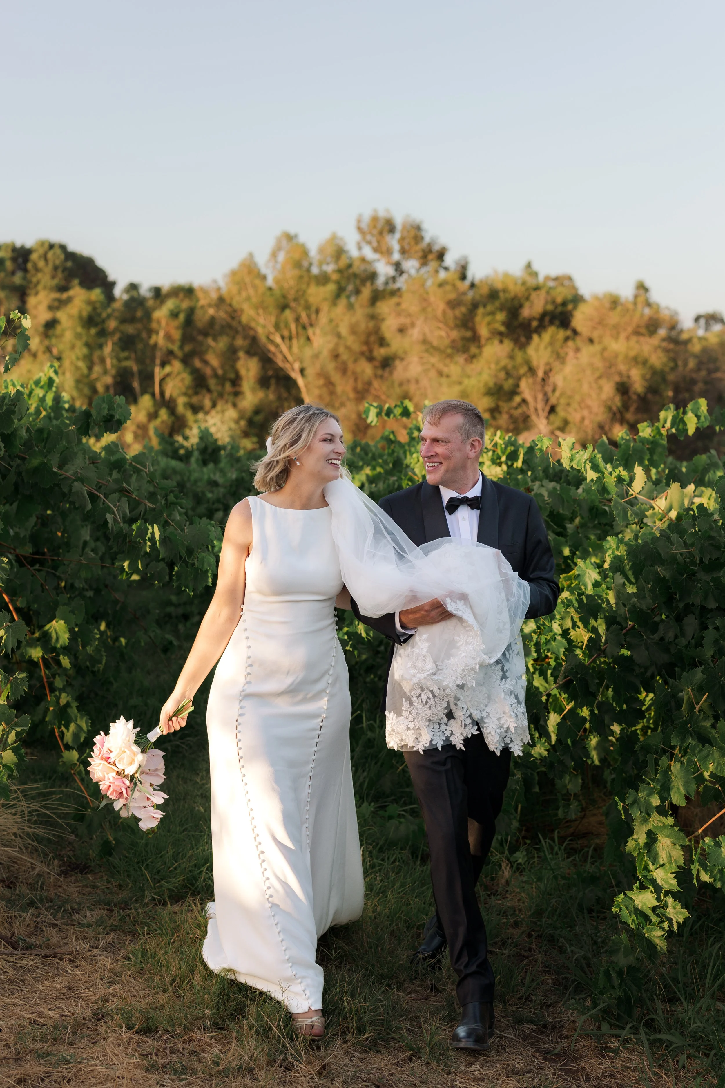 A newlywed couple walking through a vineyard, the bride holding a bouquet of flowers, both smiling and looking at each other.