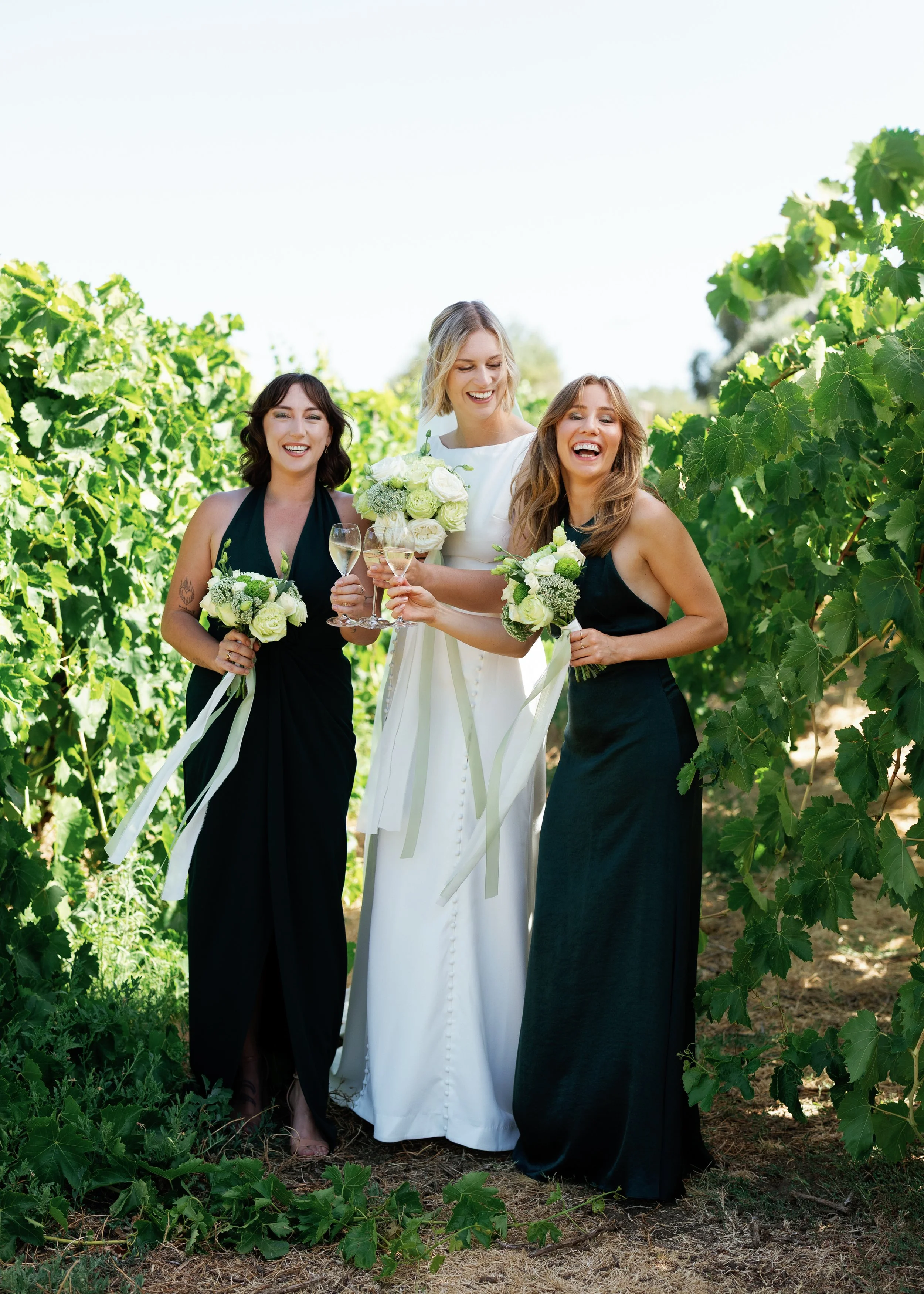 A bride and two bridesmaids celebrating outdoors in a vineyard, holding bouquets and glasses of champagne, smiling and laughing.
