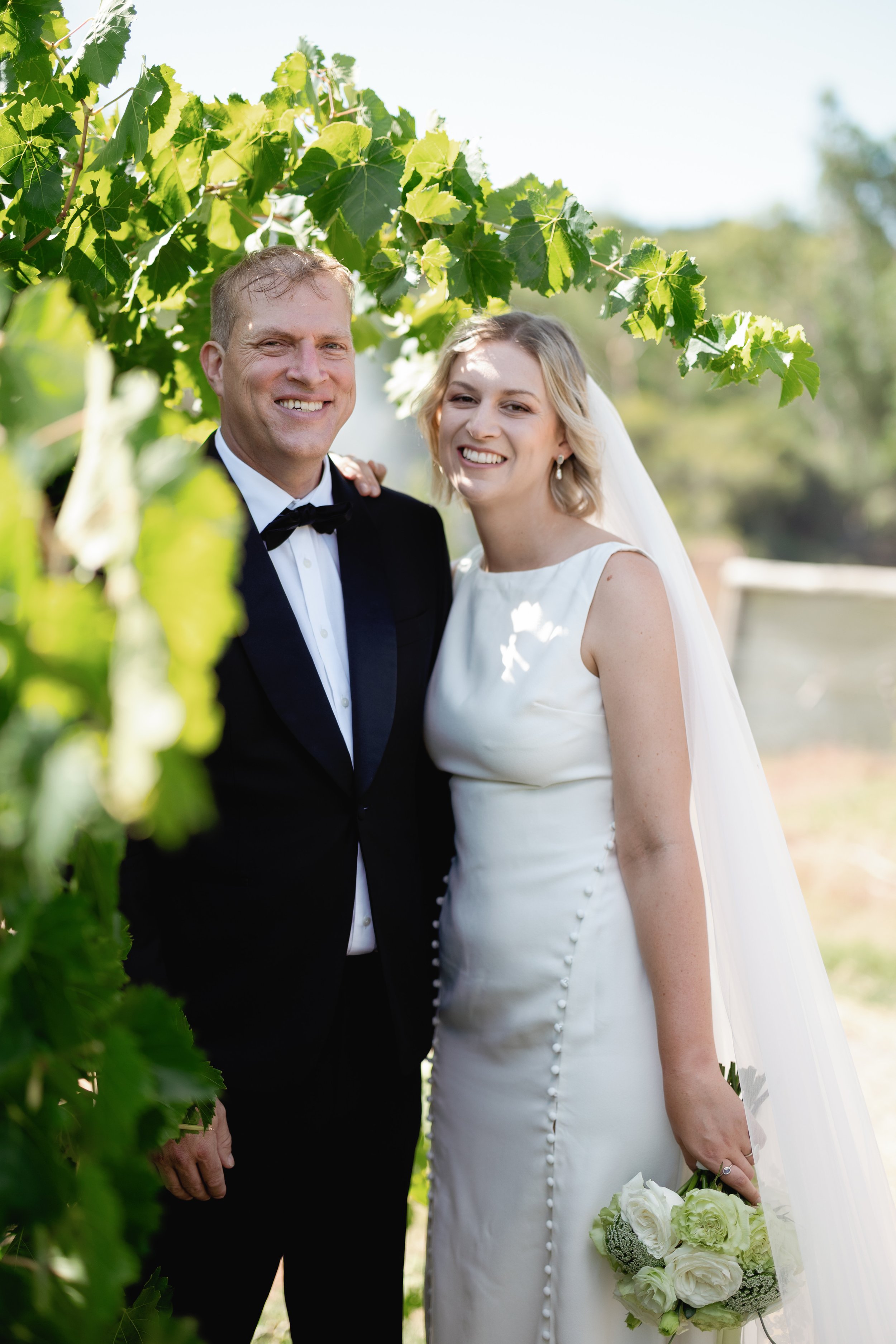 Bride and groom standing together outdoors, smiling, with lush green foliage in the background. The bride is wearing a white wedding dress and holding a bouquet of white flowers. The groom is wearing a black tuxedo with a bow tie.