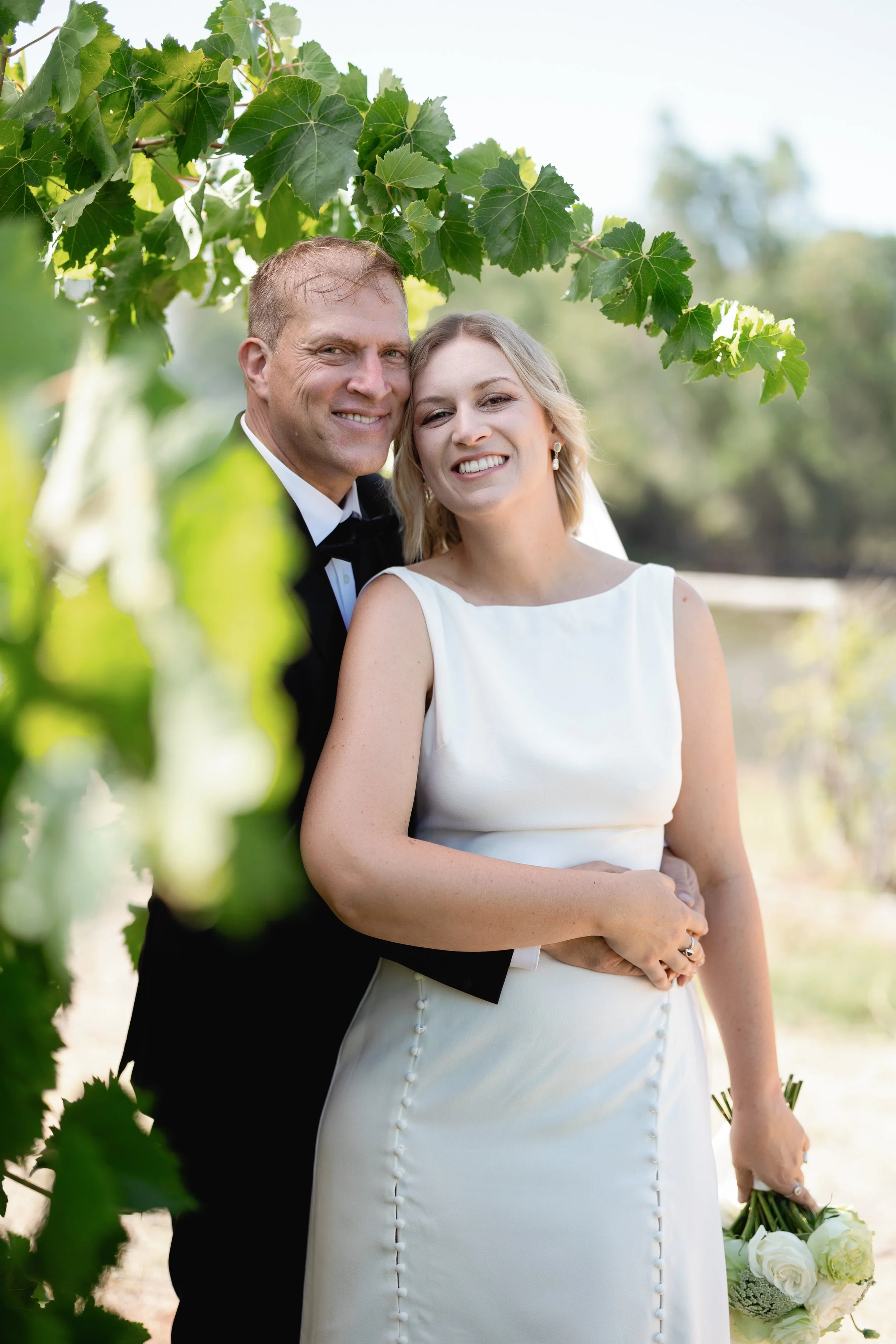 A newlywed couple in wedding attire posing outdoors among green foliage, smiling at the camera. The bride is holding a bouquet of white flowers.