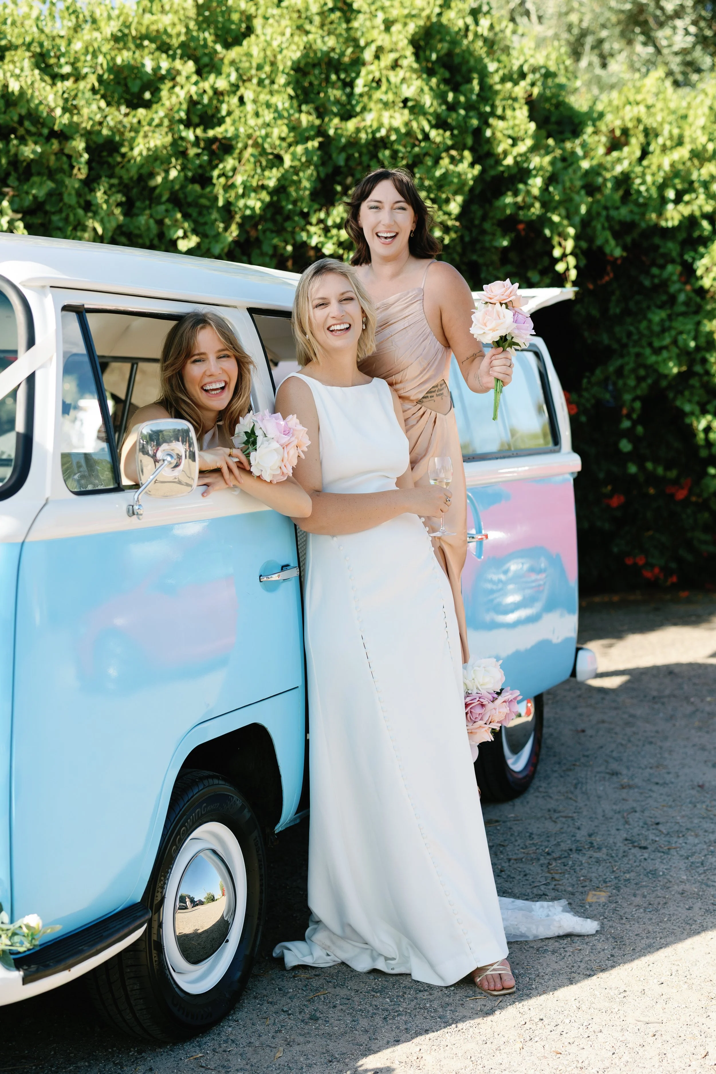 Three women celebrating, one in a white wedding dress, standing outside next to a vintage blue and white van, holding bouquets and glasses of wine, smiling and laughing under green trees.