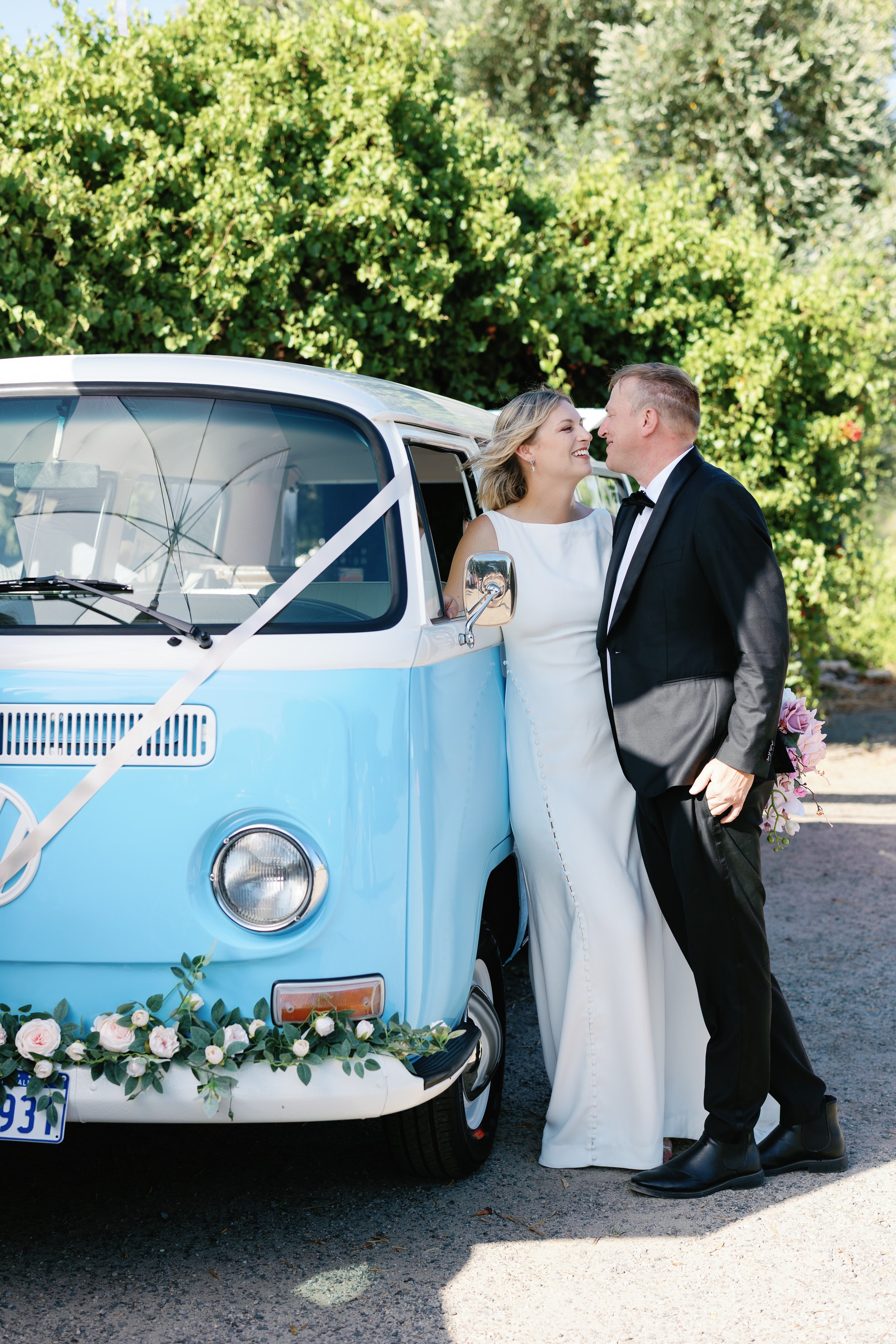 A bride and groom in wedding attire are standing next to a vintage blue and white Volkswagen van decorated with flowers and ribbons, outdoors with lush green trees in the background.