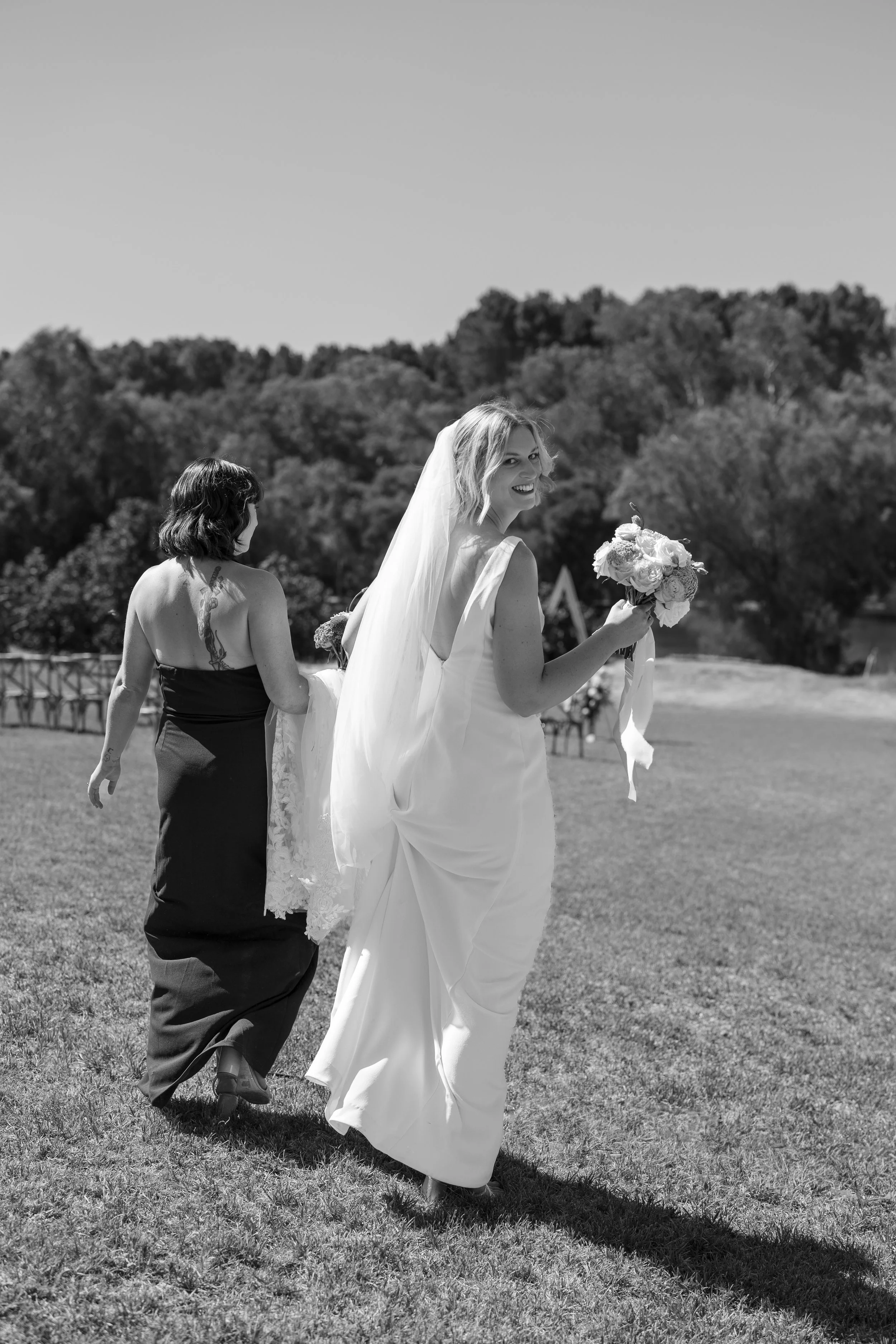 A bride in a wedding dress holding a bouquet, smiling as she walks outdoors on a sunny day. A bridesmaid in a dark dress walks behind her, adjusting the bride's dress, on a grassy field with trees in the background.