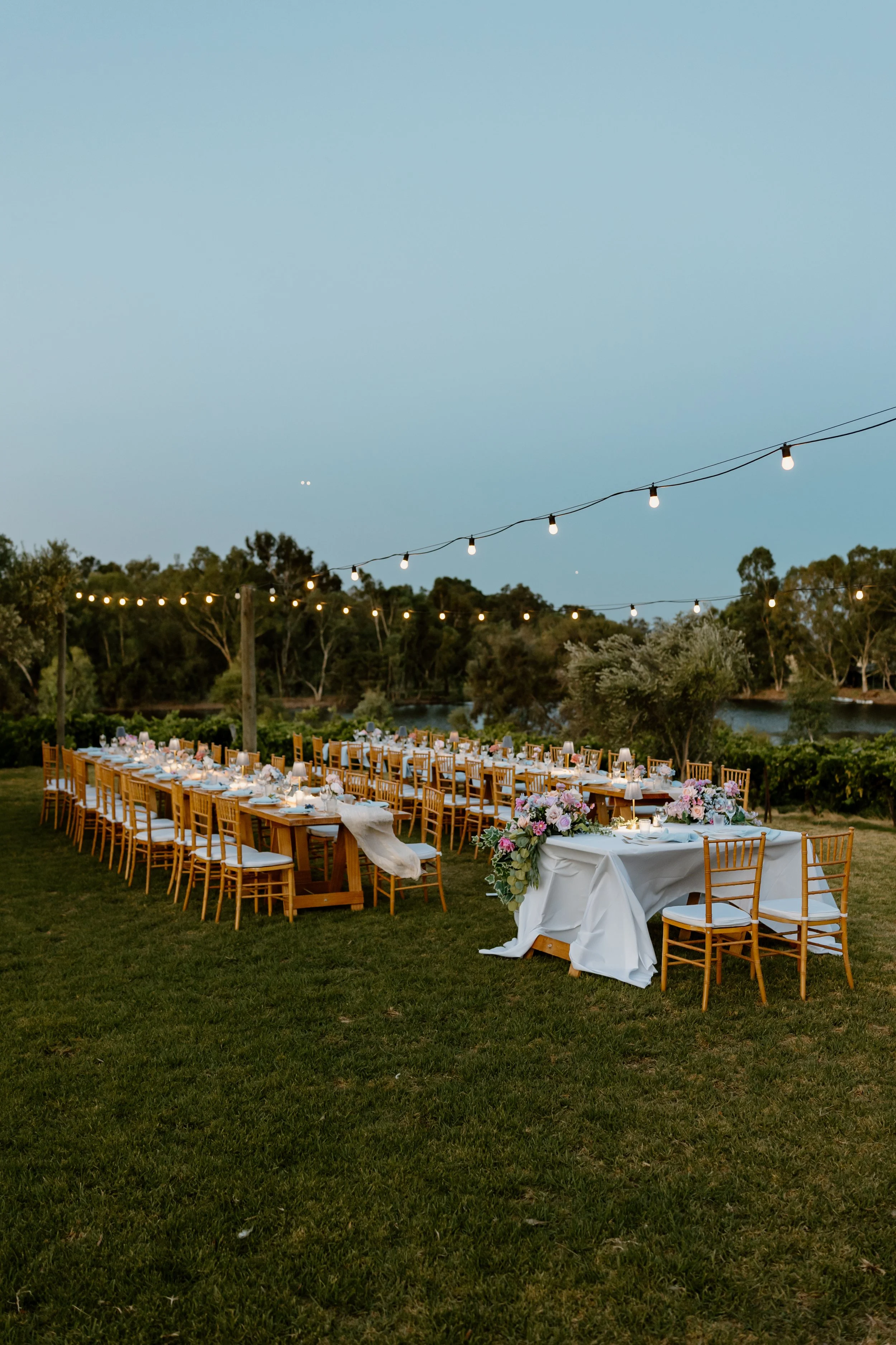Outdoor wedding reception setup with long wooden tables, white tablecloths, floral centerpieces, and string lights, overlooking a river and surrounded by trees at dusk.