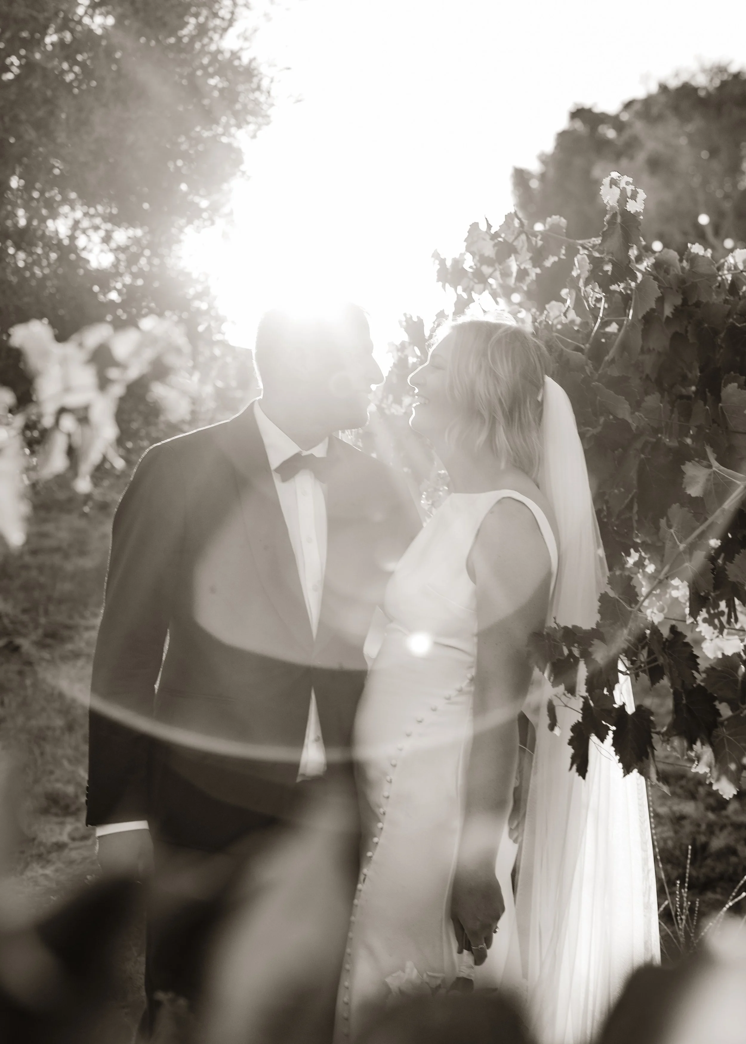 A black-and-white photo of a bride and groom standing close together outdoors, facing each other with noses nearly touching, with sunlight shining behind them.