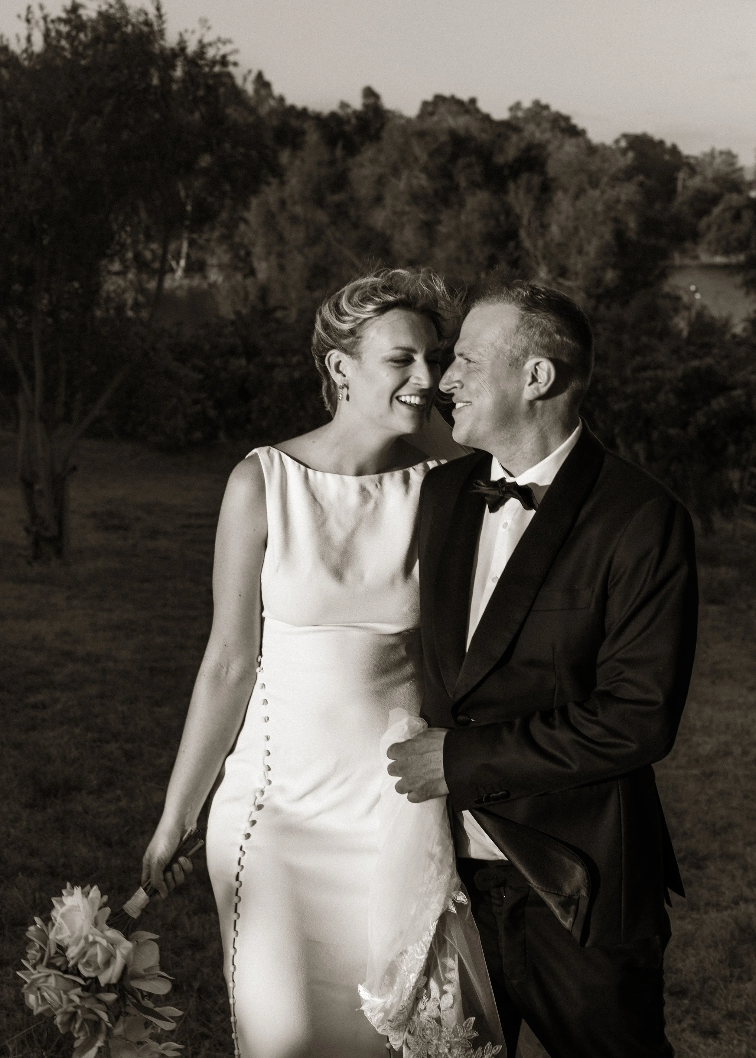 Black and white photo of a bride and groom smiling at each other outdoors, with trees and hills in the background.