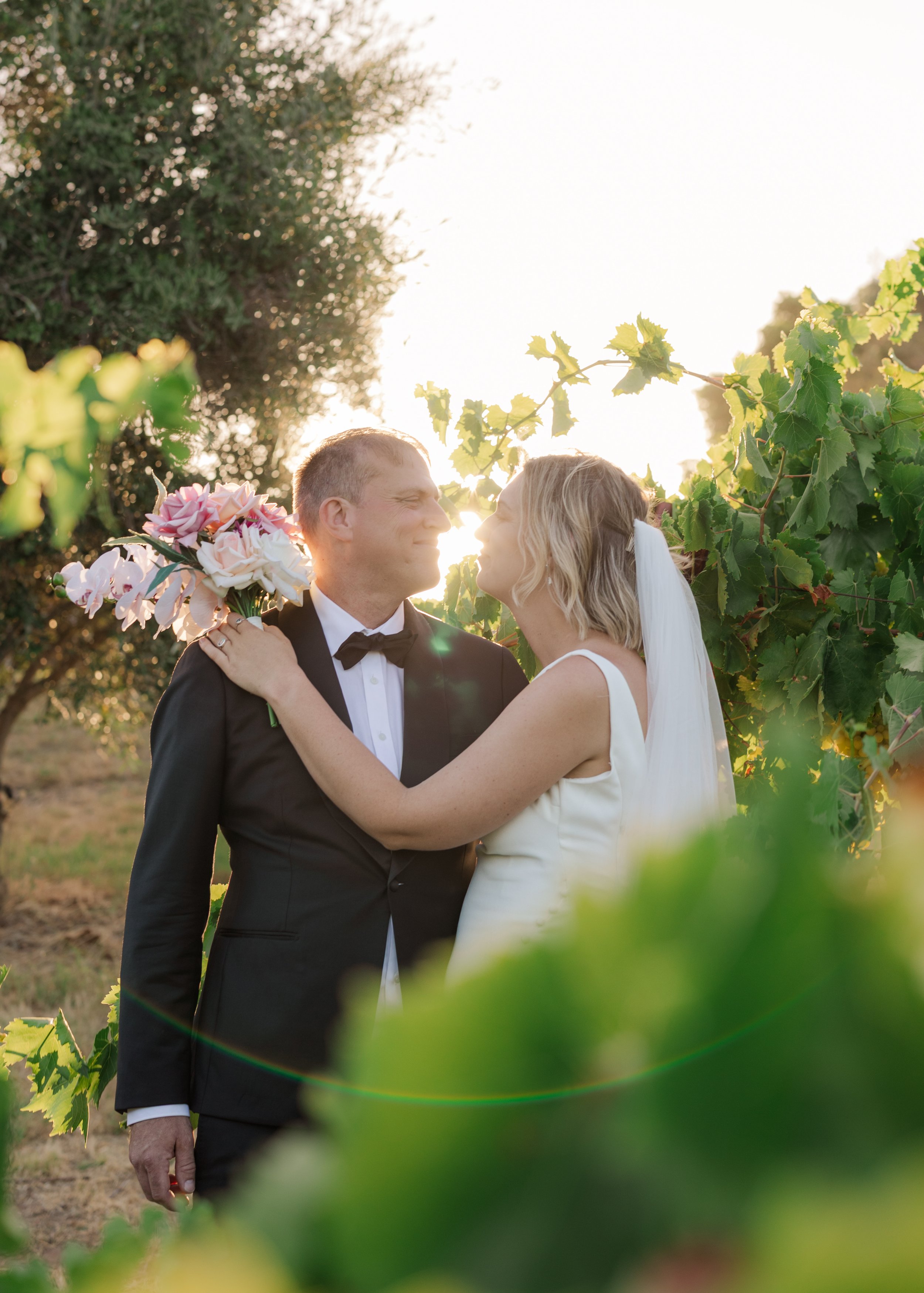 A bride and groom celebrating their wedding outdoors during sunset, with the bride holding a bouquet of pink and white flowers, surrounded by green vineyard vines.