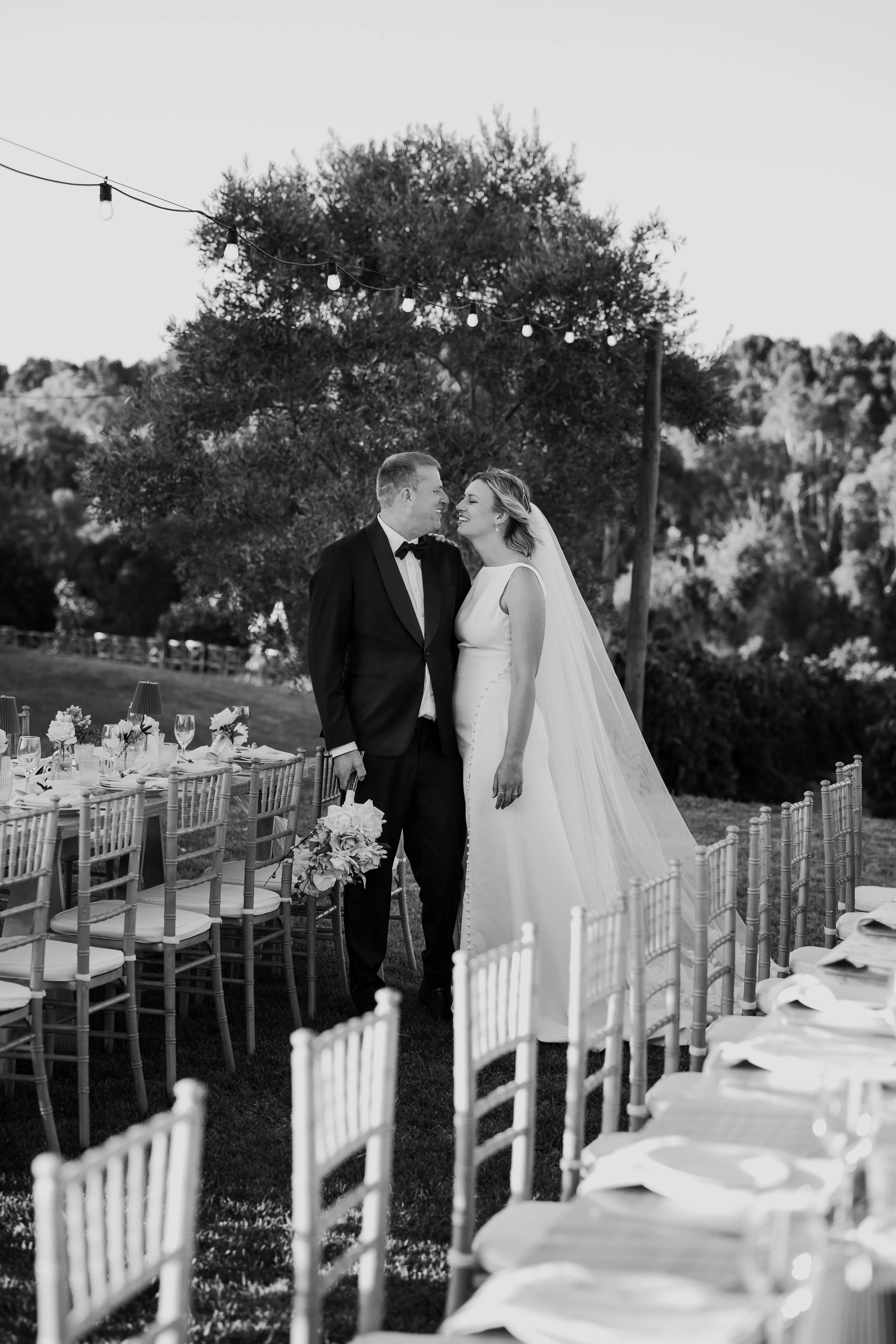 Black and white photo of a bride and groom standing close together outdoors, smiling and looking at each other, surrounded by decorated tables and chairs at a wedding reception.