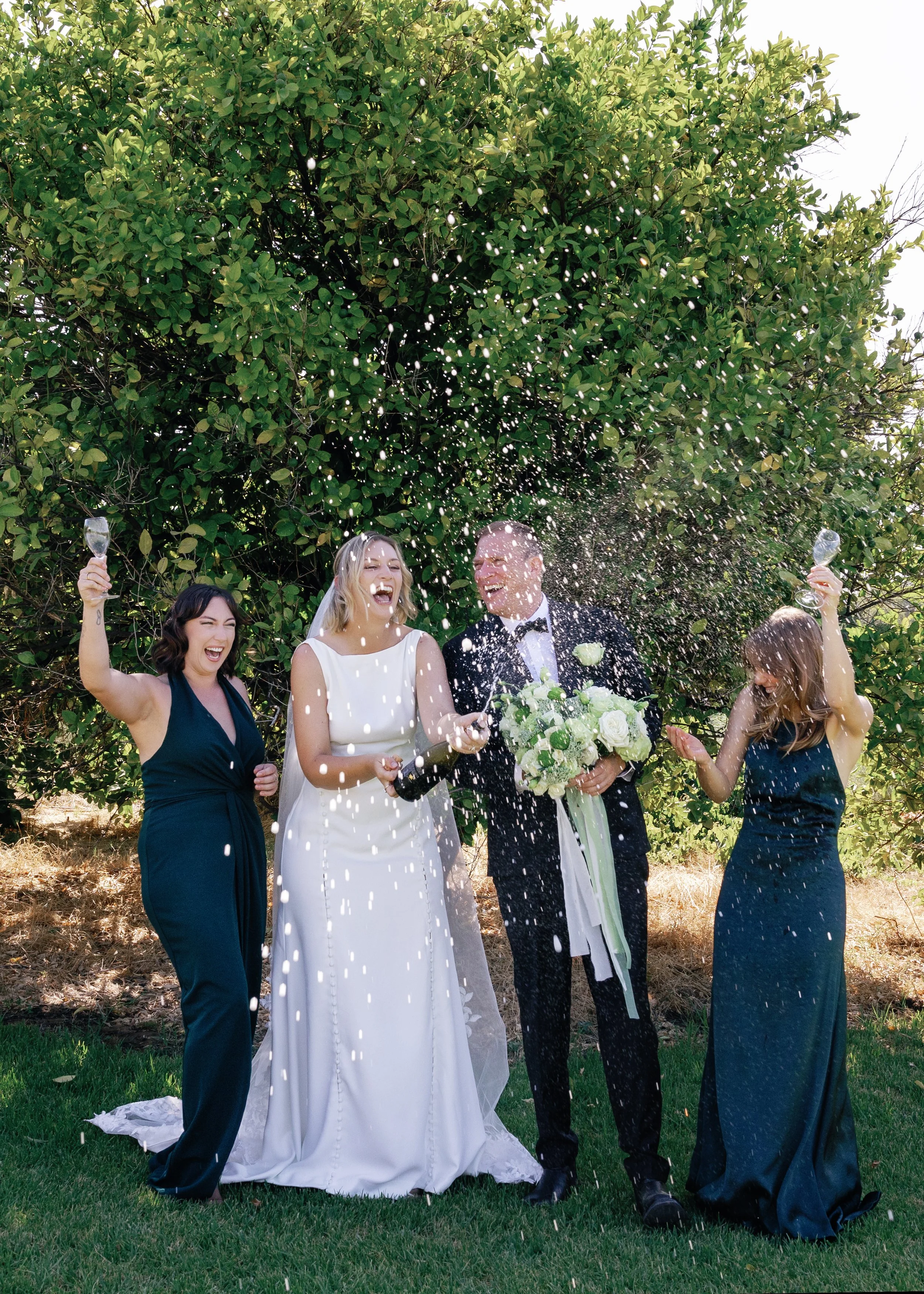 A wedding celebration with four people outdoors, two women and two men, one wearing a bride's wedding dress and veil, holding a champagne bottle, and the other man in a tuxedo holding a bouquet of white flowers. The parties are spraying champagne and
