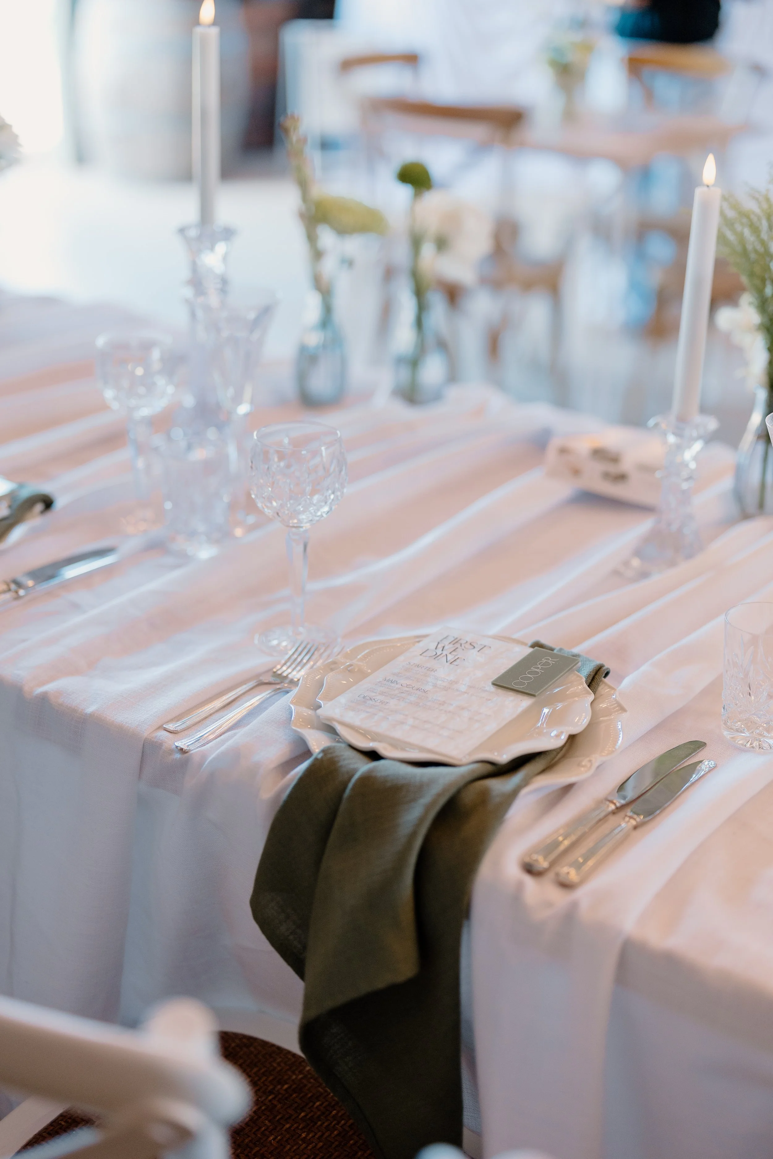 Elegant dining table set with white tablecloth, crystal glassware, tall candles, floral centerpieces, and place settings, likely for a wedding or special event.