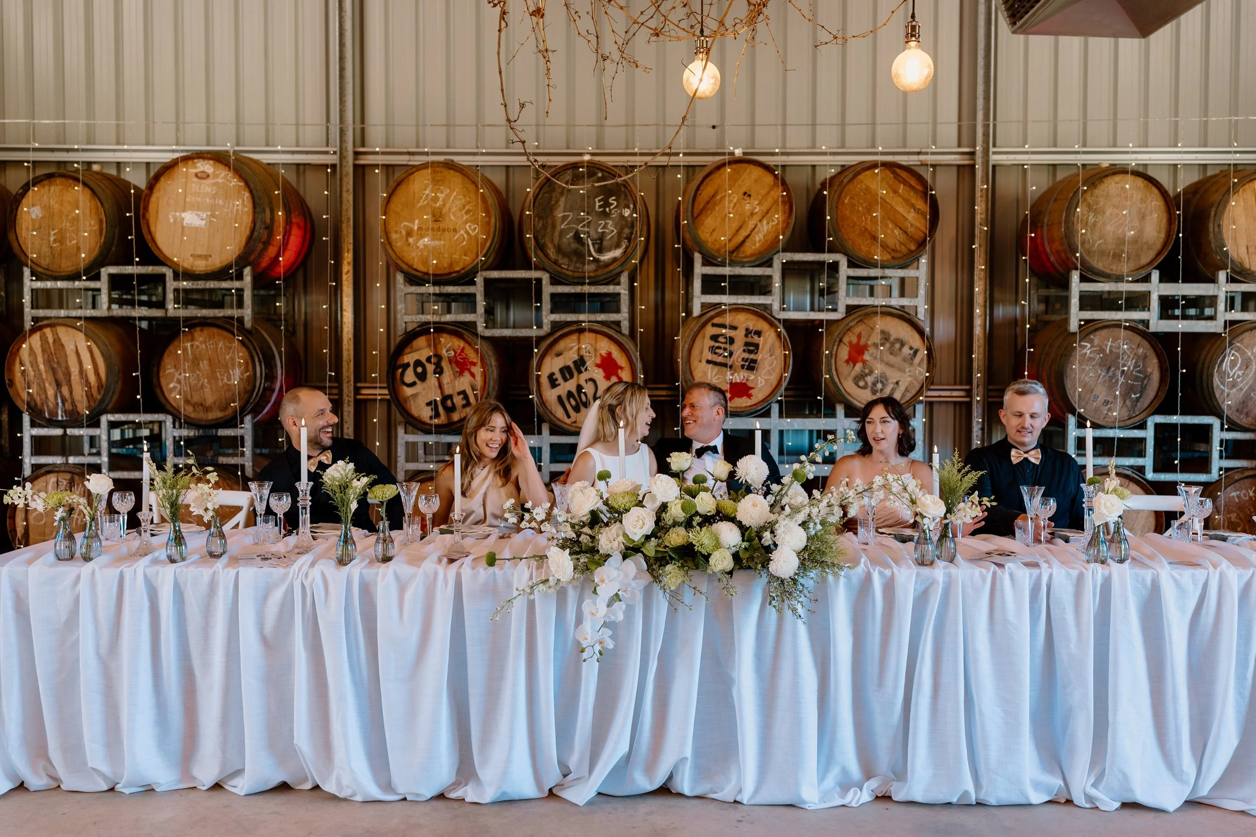 Wedding reception with a long table decorated with a white tablecloth, floral centerpiece, and candles, with six people seated against a backdrop of large wine barrels and string lights.