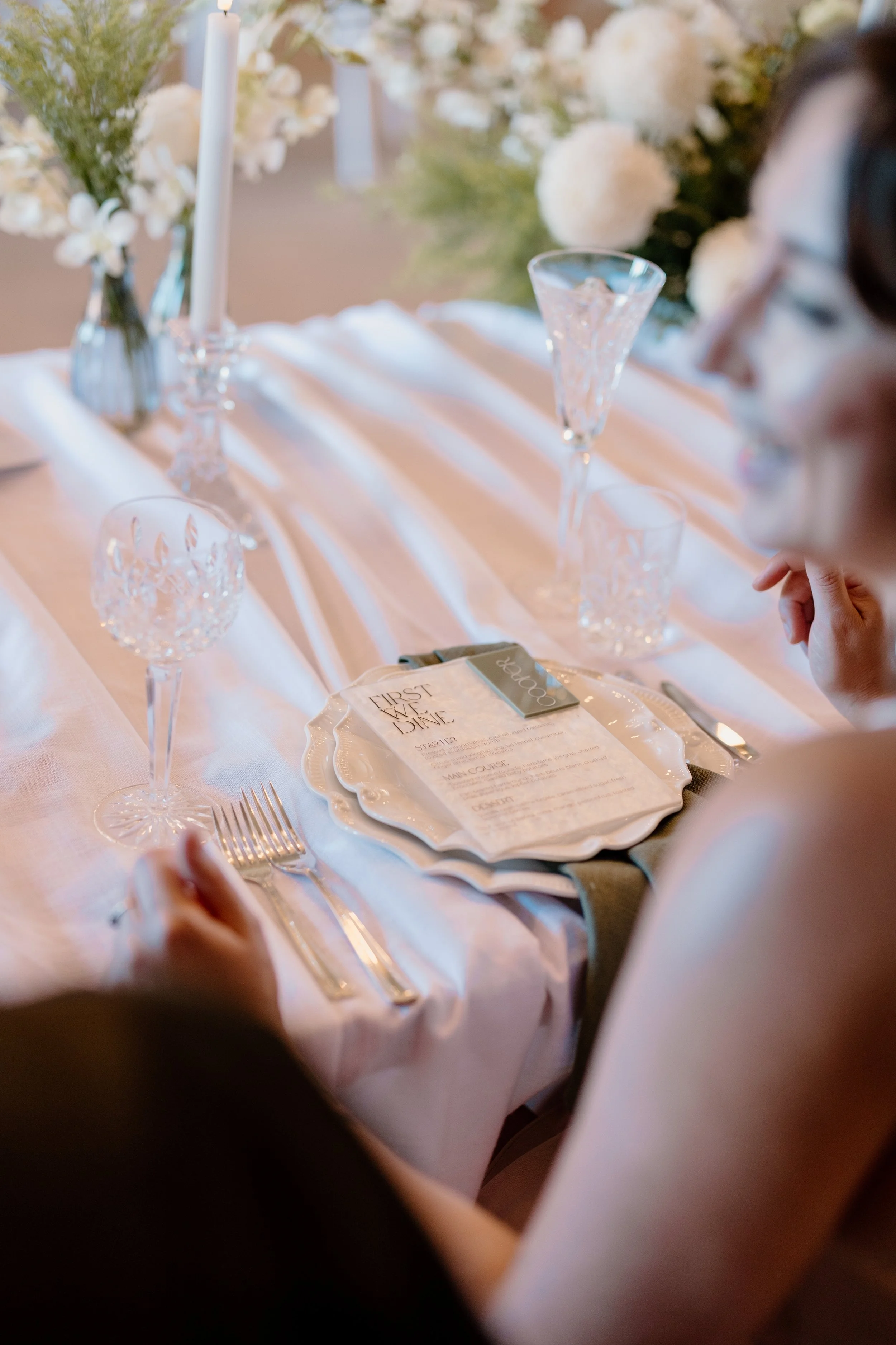 Elegant wedding reception table with white tablecloth, crystal glasses, cutlery, floral centerpiece, and a menu card.