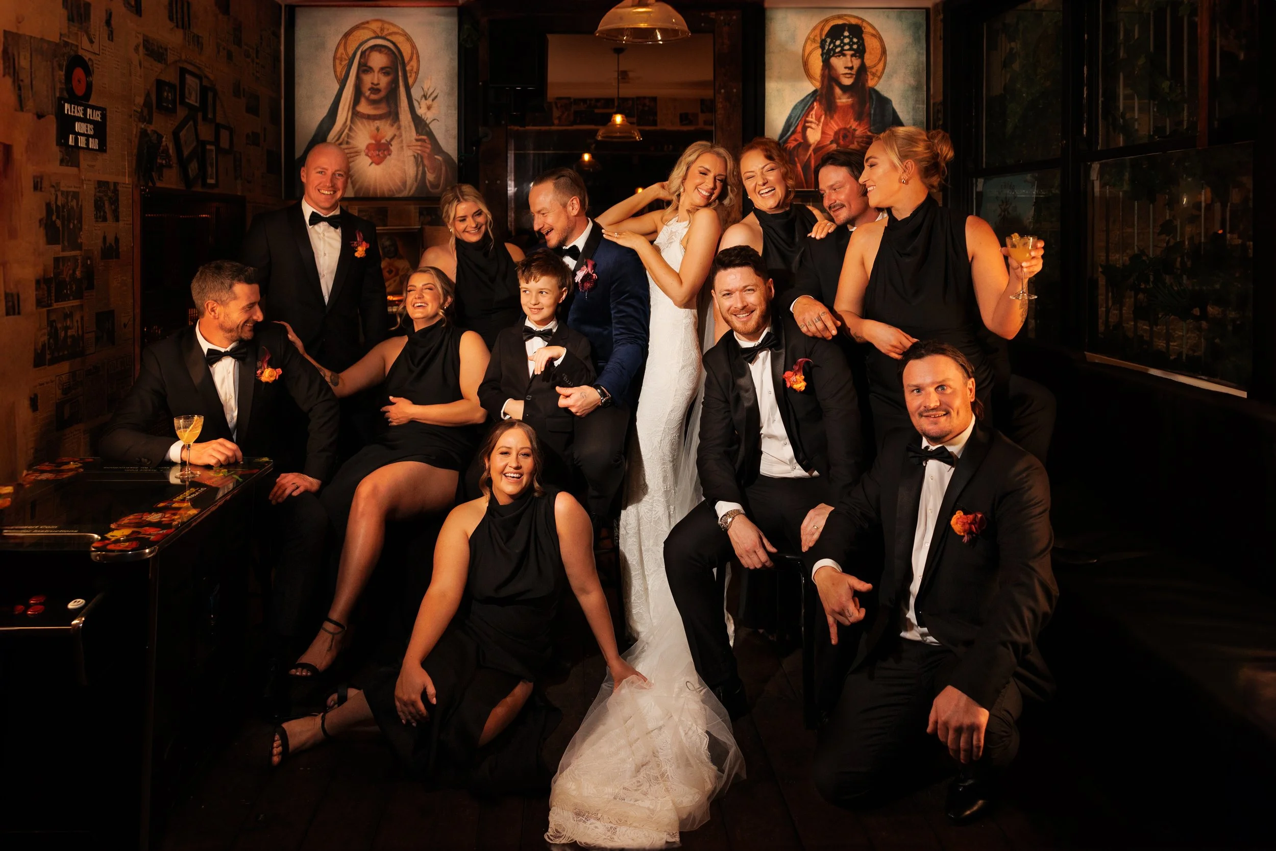 Group of people at a wedding celebration, including the bride in a white gown, surrounded by friends and family dressed in formal attire, inside a dimly lit venue decorated with religious artwork and vintage posters.