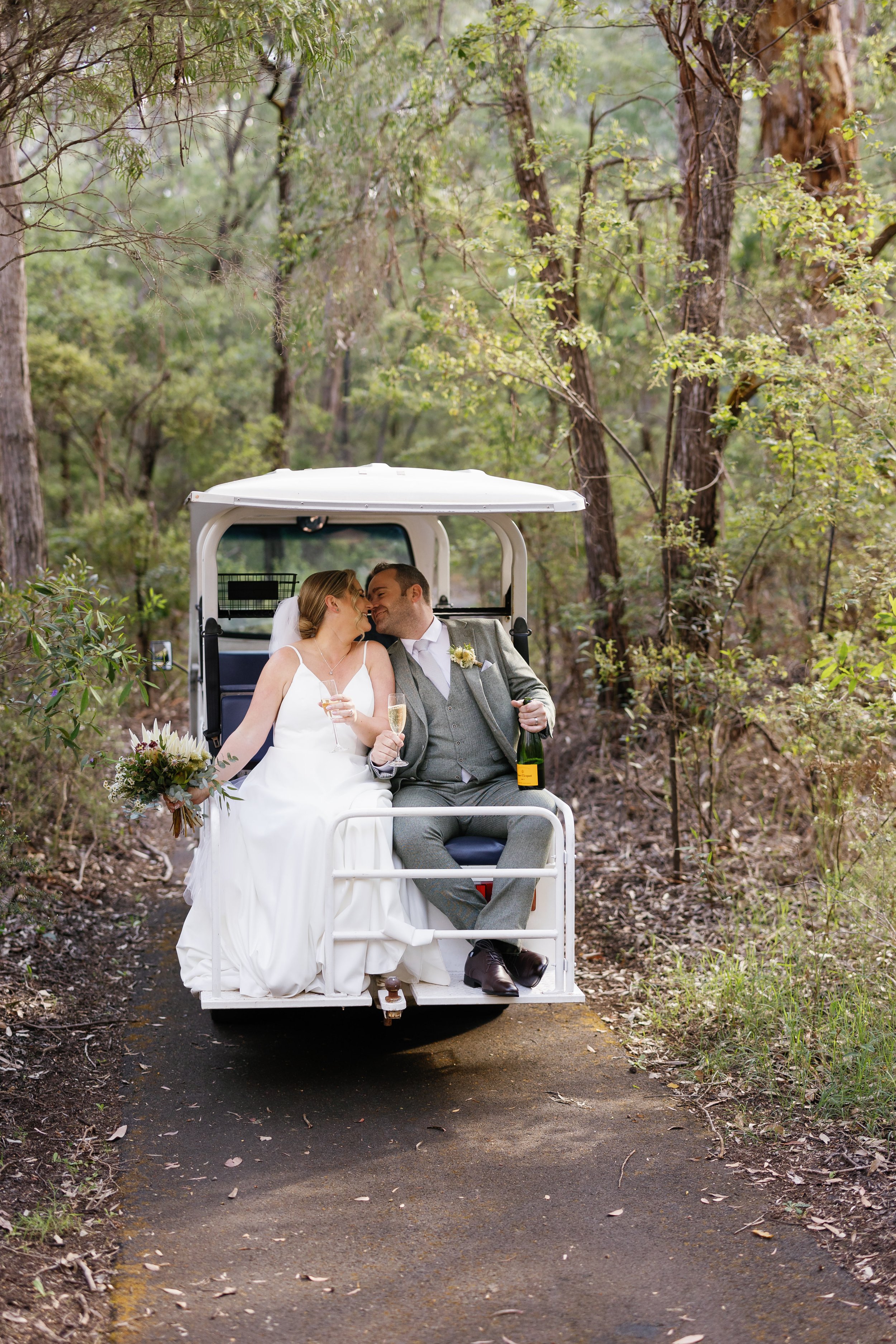 A newlywed couple in wedding attire sharing a kiss on a golf cart in a wooded area, with the bride holding a bouquet and the groom holding a bottle of champagne.