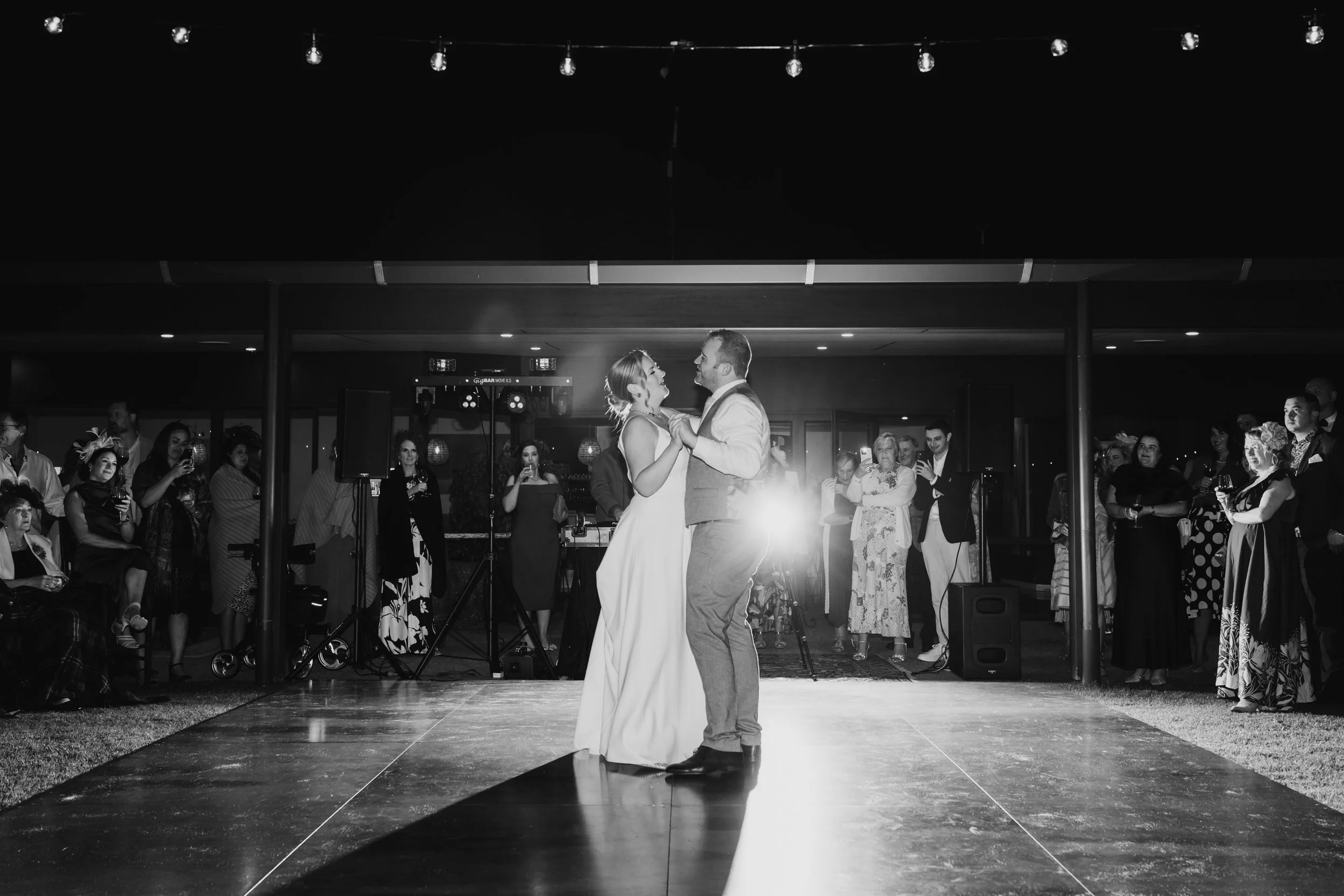 A bride and groom share a dance during their wedding reception on a dark dance floor, surrounded by guests watching and taking photos.