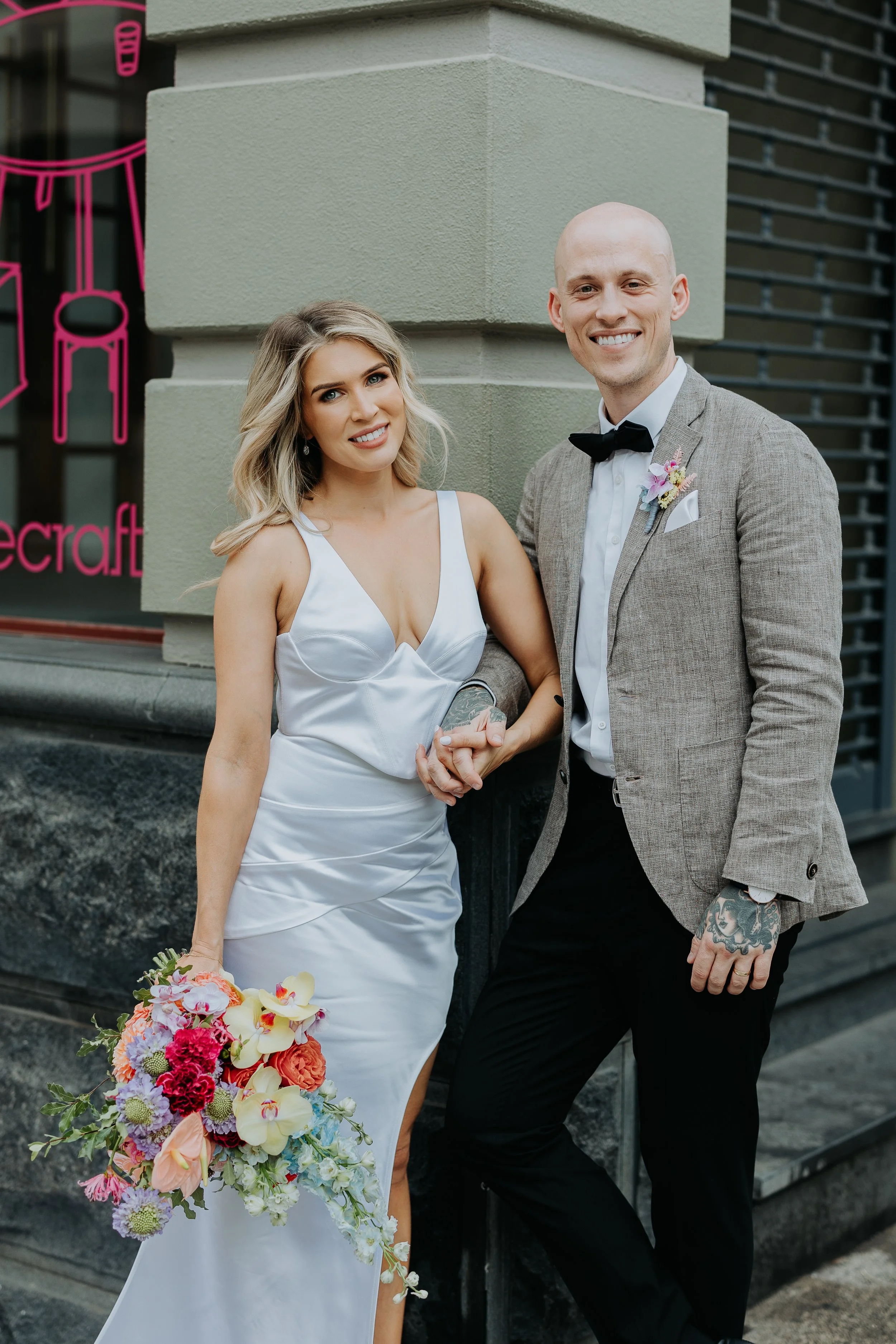 A bride and groom standing outside a building, holding hands, smiling, dressed in wedding attire. The bride wears a white satin wedding dress holding a colorful bouquet, and the groom wears a gray blazer, black bow tie, and has tattoos on his hand.